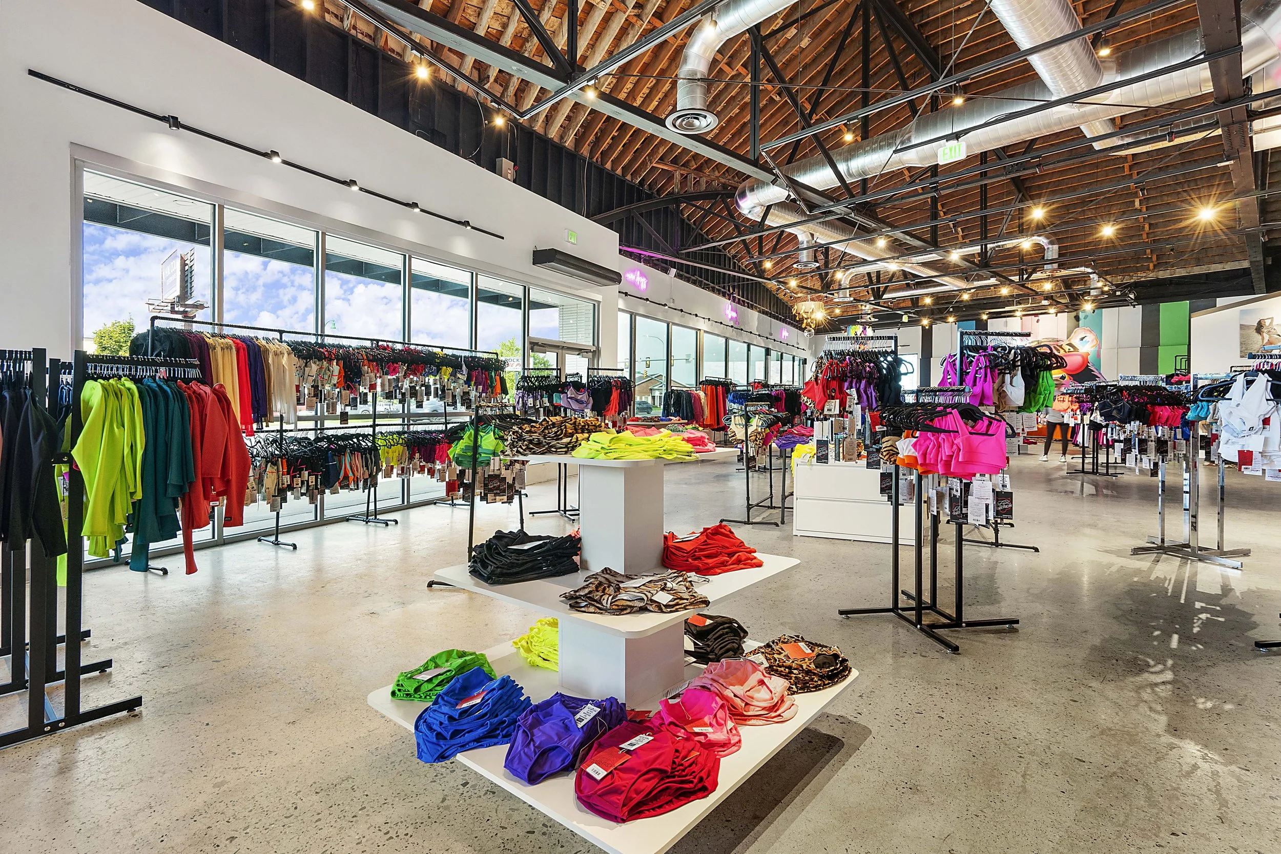 Inside a retail store selling colorful athletic wear and swimwear, with clothing racks and tables displaying various clothing items. Large windows let in natural light, and the high ceiling has exposed industrial pipes and lighting.