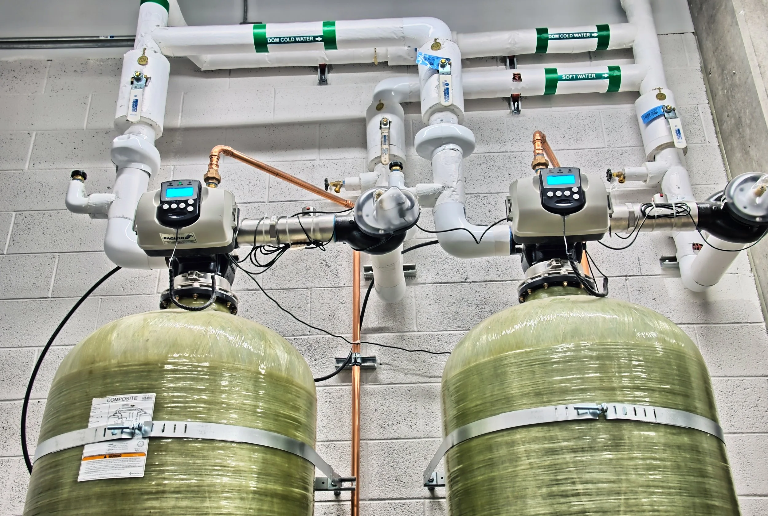 Indoor water treatment or filtration system with two large green tanks connected to white piping and electronic control units mounted above each tank.