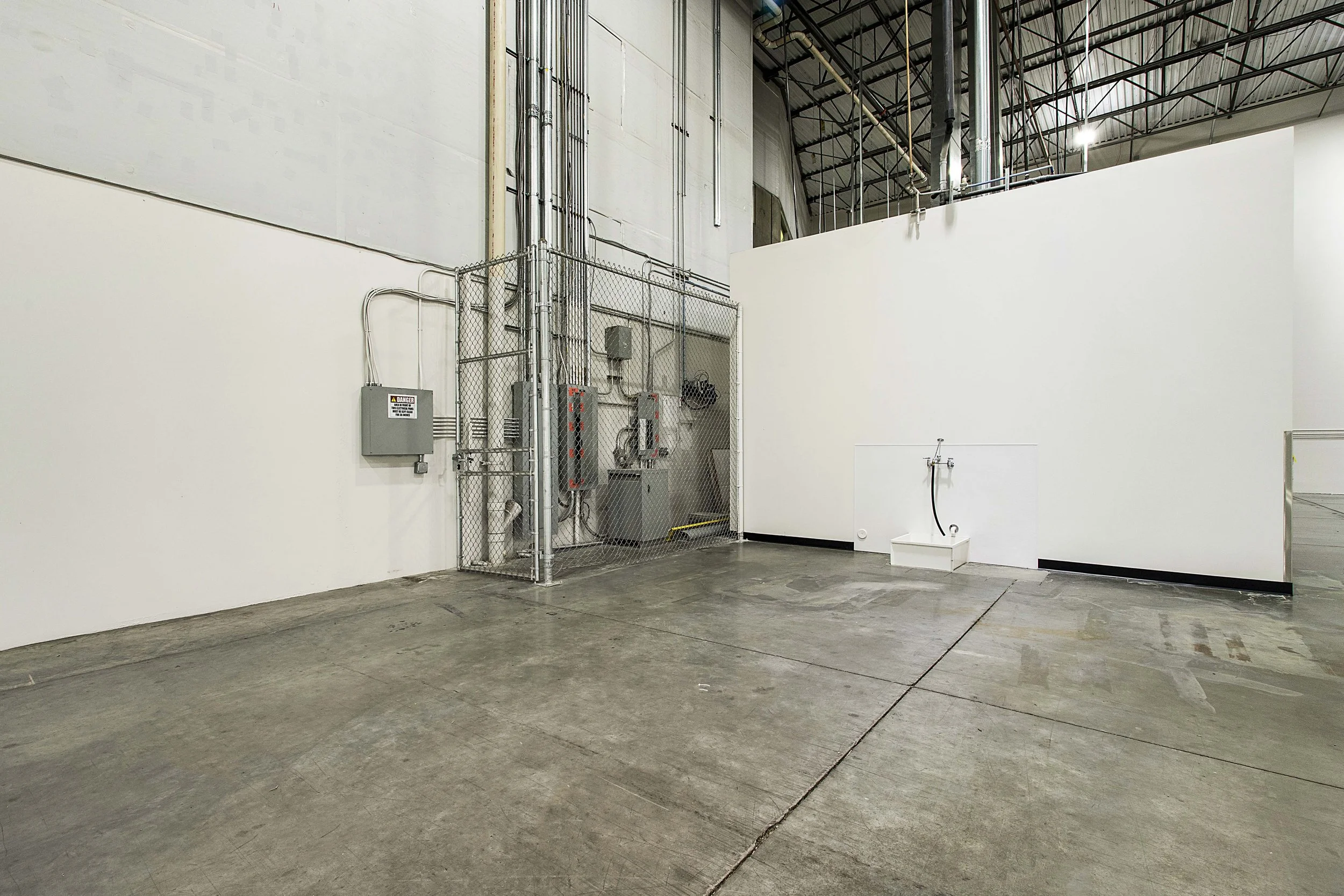 Empty industrial warehouse with a white wall, concrete floor, and visible electrical and plumbing fixtures.