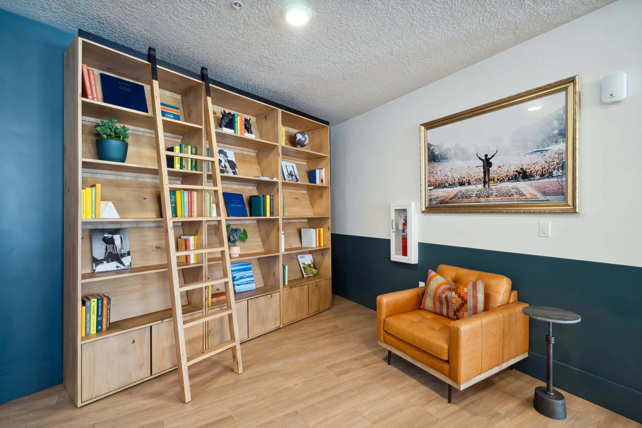 A cozy room with a large wooden bookshelf, a ladder, a tan leather armchair with a patterned pillow, a small round side table, a framed artwork of a person with raised arms, and a fire safety box on the wall.