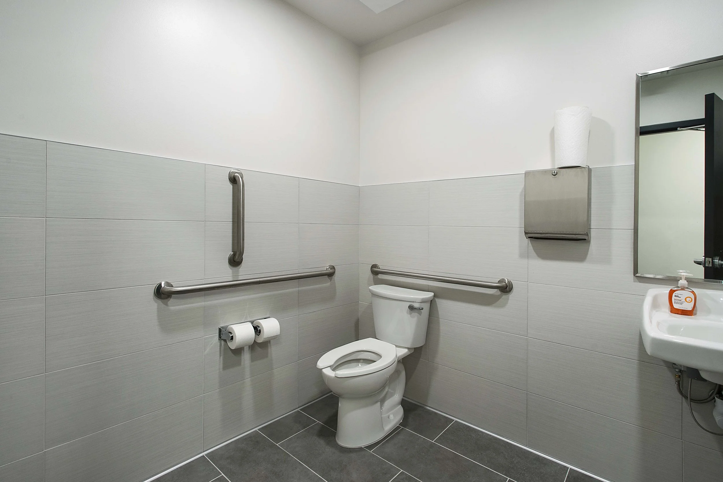 Accessible bathroom with a toilet, horizontal and vertical grab bars, toilet paper rolls, a mirror, paper towel dispenser, and a soap container, with tiled walls and floor.