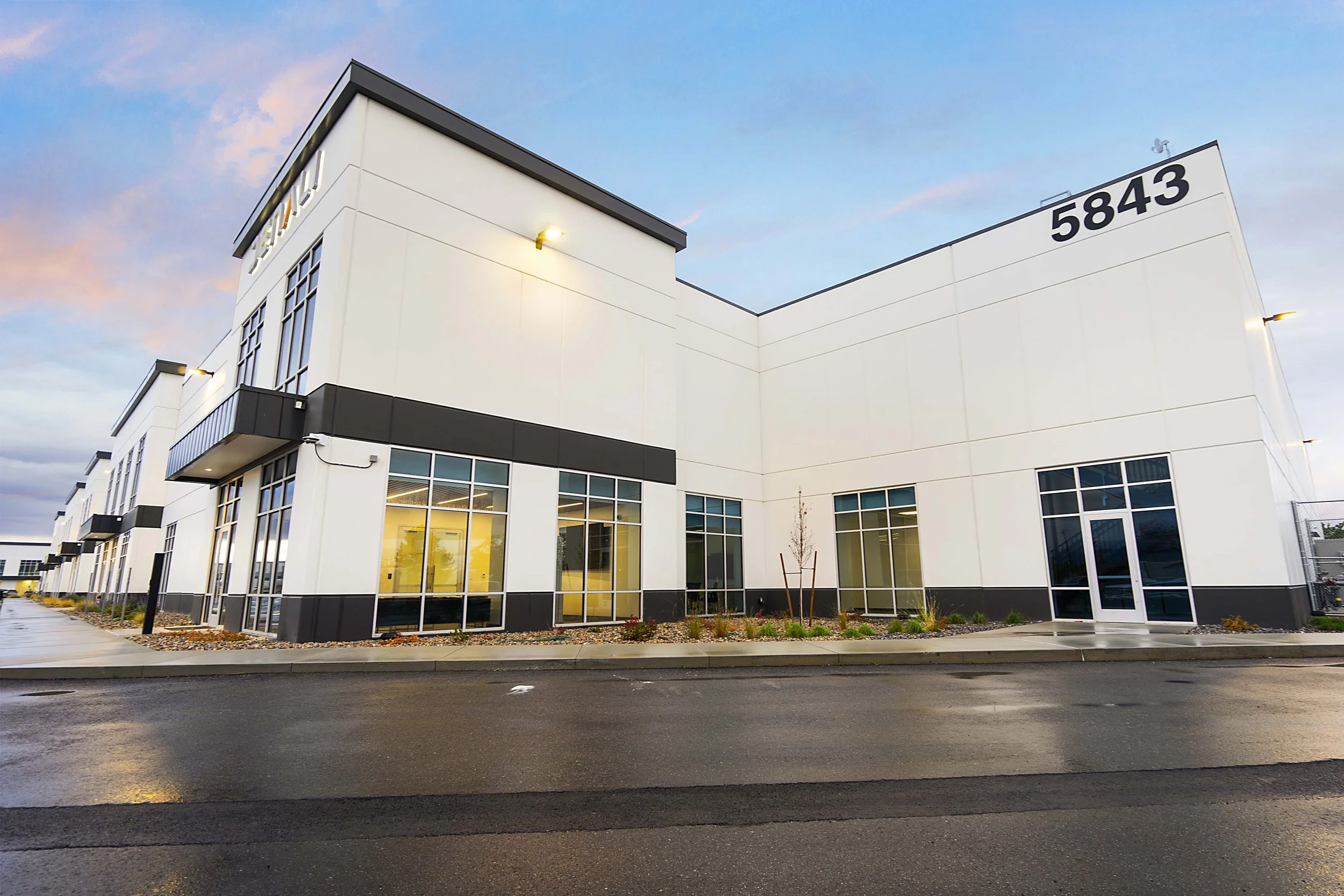 Exterior of a modern white commercial building with large glass windows, black trim, and a street in front, under a sky with pink clouds.