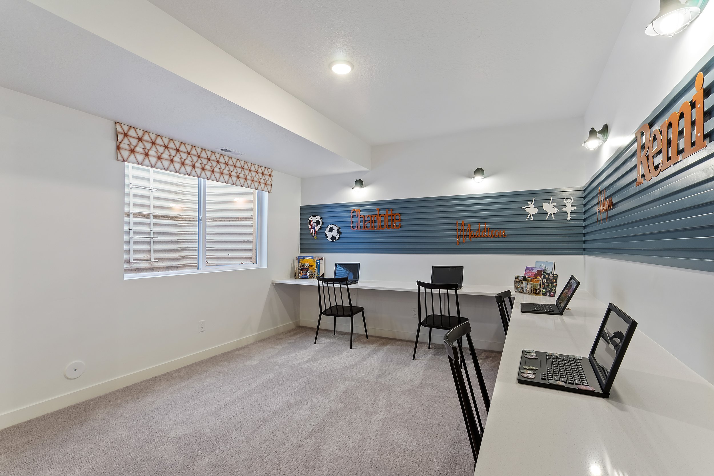 A gaming or activity room with a white counter along the wall equipped with four laptops, black chairs, a window with a patterned valance, and a wall decorated with words, pictures, and game-themed decor.