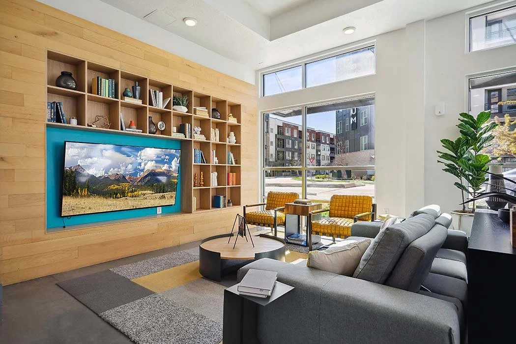 Modern living room with a gray sectional sofa, yellow armchairs, and a large window showing an urban view. Wooden feature wall with mounted flat-screen TV and a built-in bookshelf.