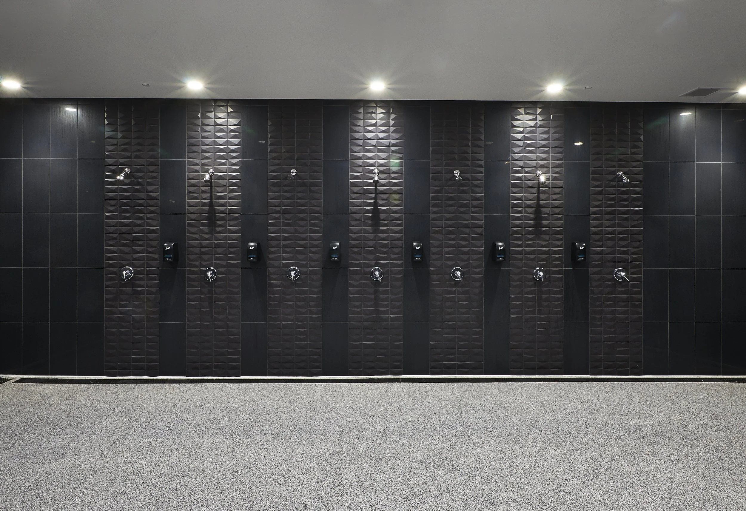 A row of shower heads and soap dispensers on a black tiled wall in a modern shower room.