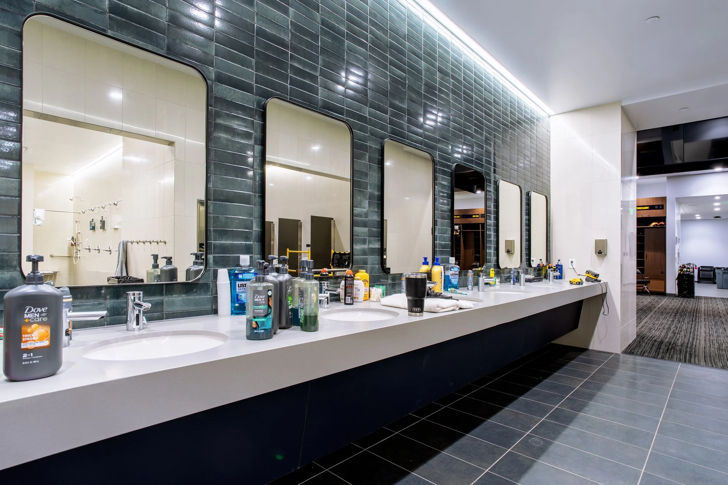 Bathroom with seven mirrors above a long white countertop with sinks, bottles of soap and personal items, dark green tiled wall, and a view into a shower area and hallway with luggage.