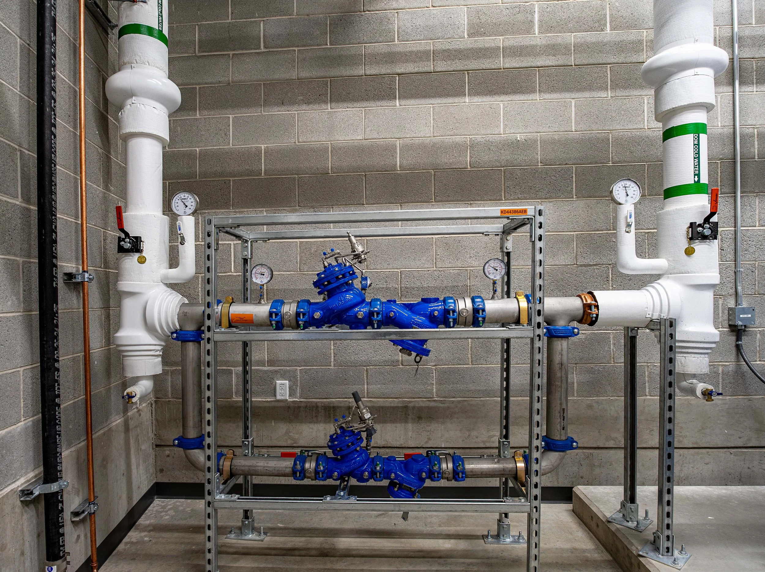 Industrial piping system with large white pipes, blue valves, pressure gauges, and metal framework mounted on a concrete wall.