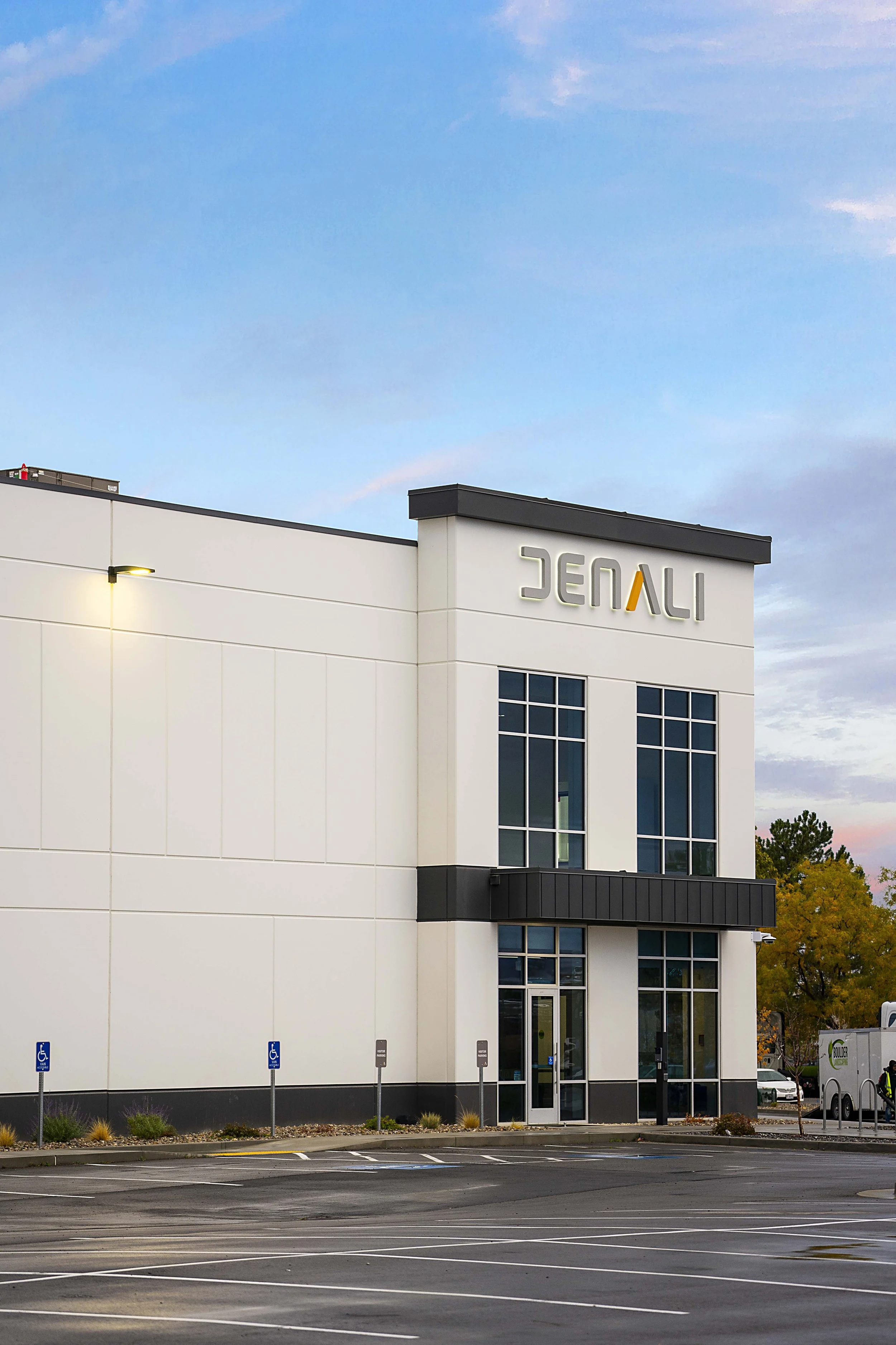 A modern, white commercial building with the name 'JENALI' on the upper part of the facade. There are large windows, a small parking lot with accessible parking spaces, and trees with autumn foliage in the background. The sky is partly cloudy with a 