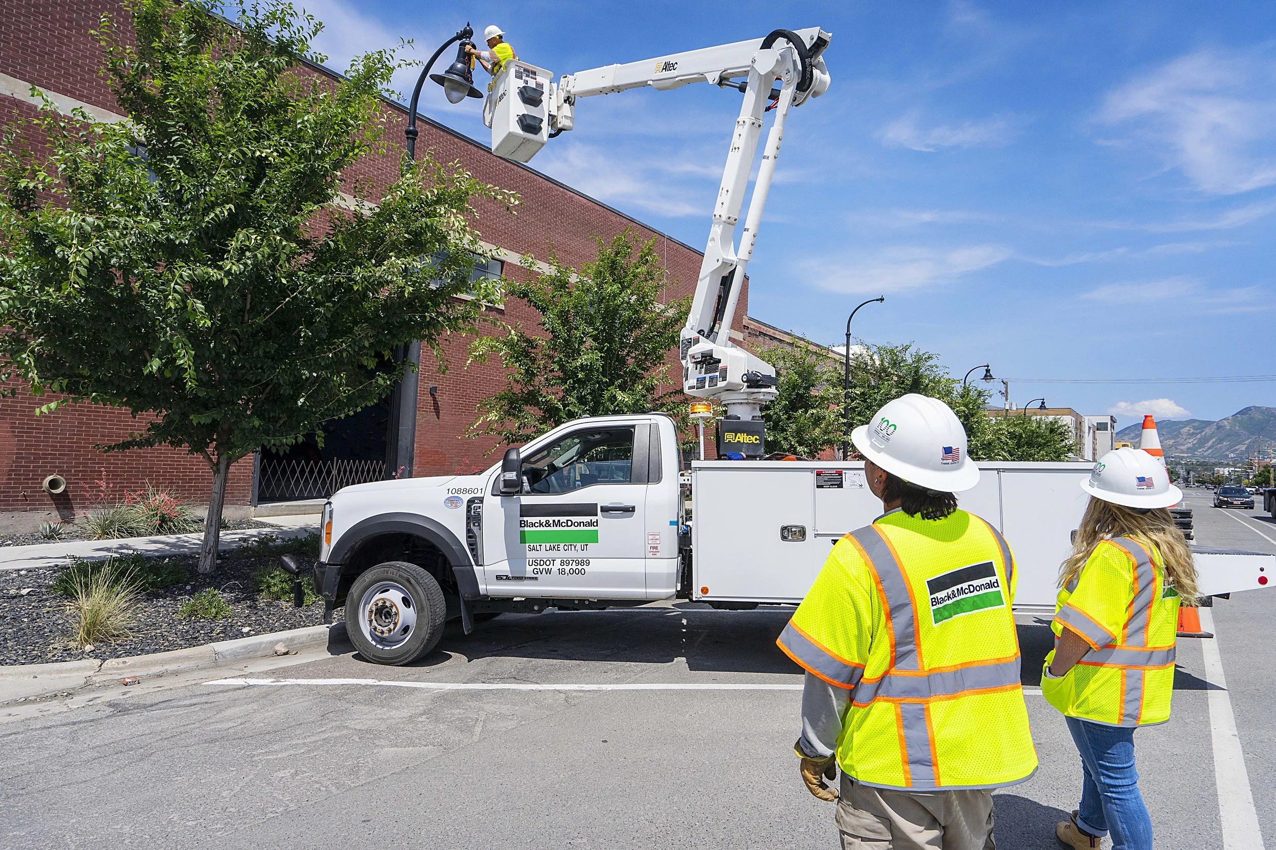 Utility workers in high-visibility safety vests and white helmets working on streetlights from a bucket truck on a city street with trees, buildings, and mountains in the background.