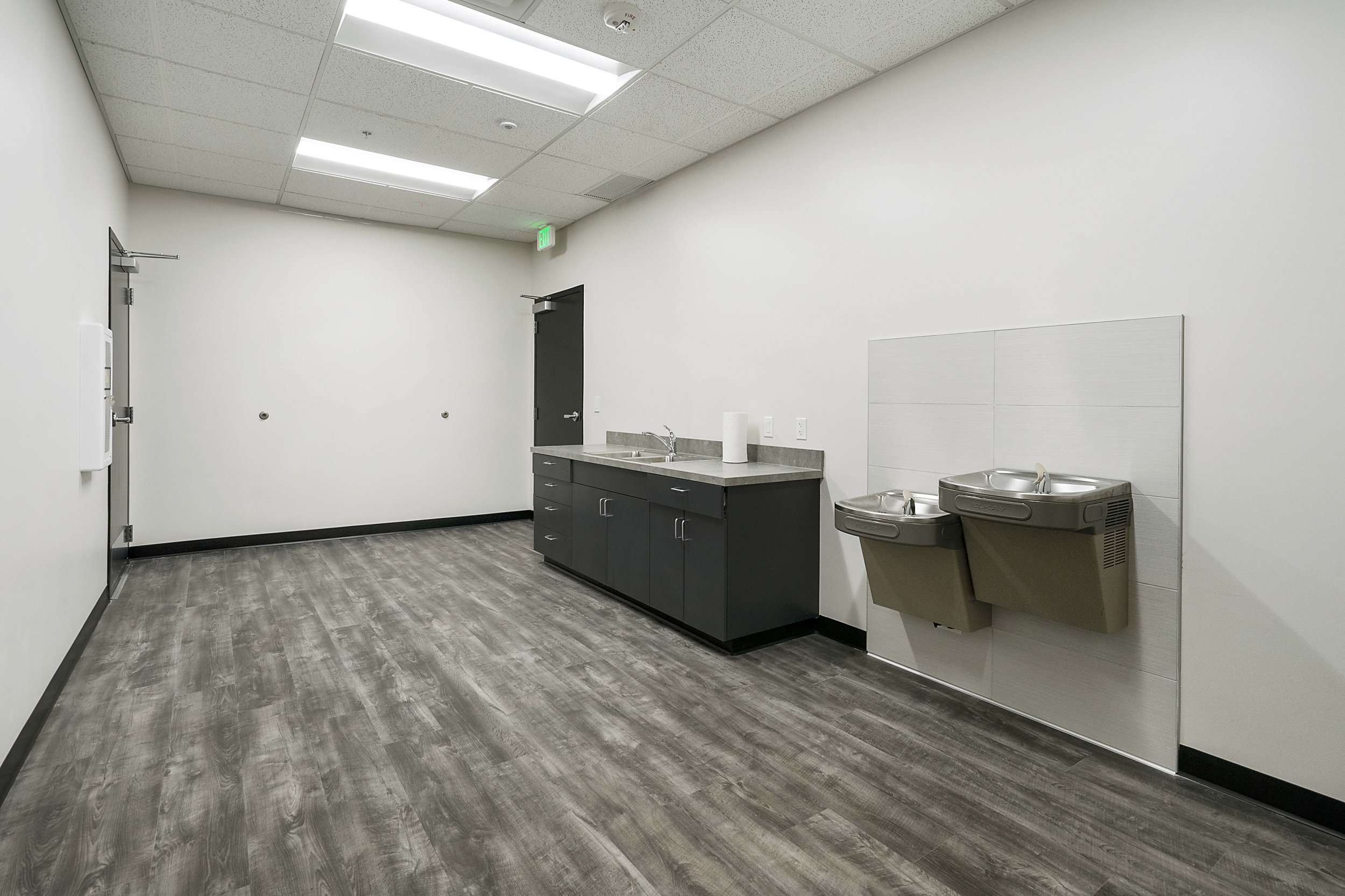 Empty break room or kitchen area with a gray countertop, sink, paper towel holder, two water fountains, and a white wall with a closed door, in a modern building.