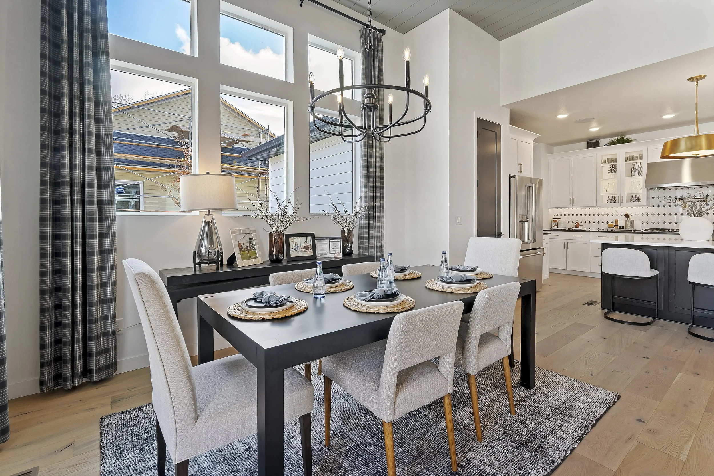 Modern dining area with a black dining table set for six, four beige upholstered chairs, a black sideboard with lamps and picture frames, large windows, and a view of the kitchen with white cabinets, black island, and stainless steel refrigerator, ch