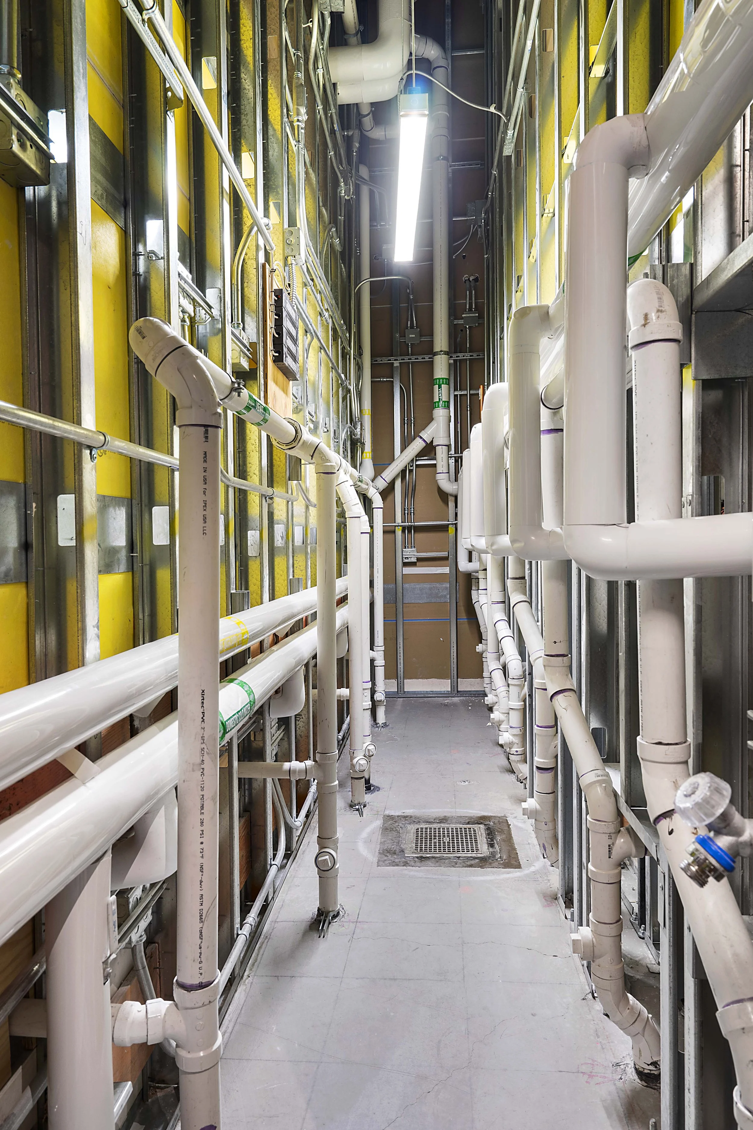 A narrow utility corridor with white pipes, metal framework, and yellow insulation panels on the walls, illuminated by fluorescent lighting.
