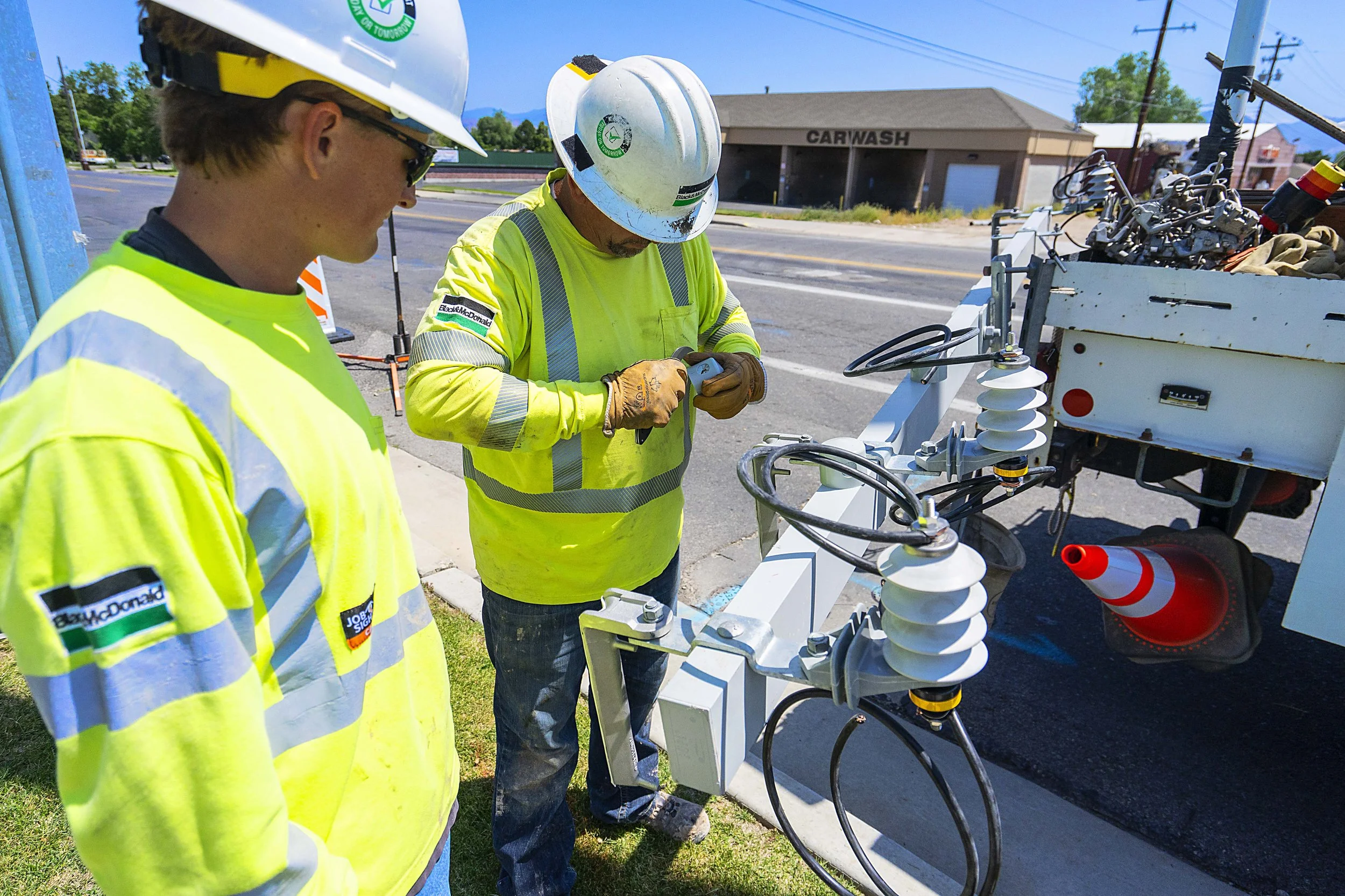 Two workers in high-visibility safety jackets and helmets inspecting or working on electrical equipment near a roadside.