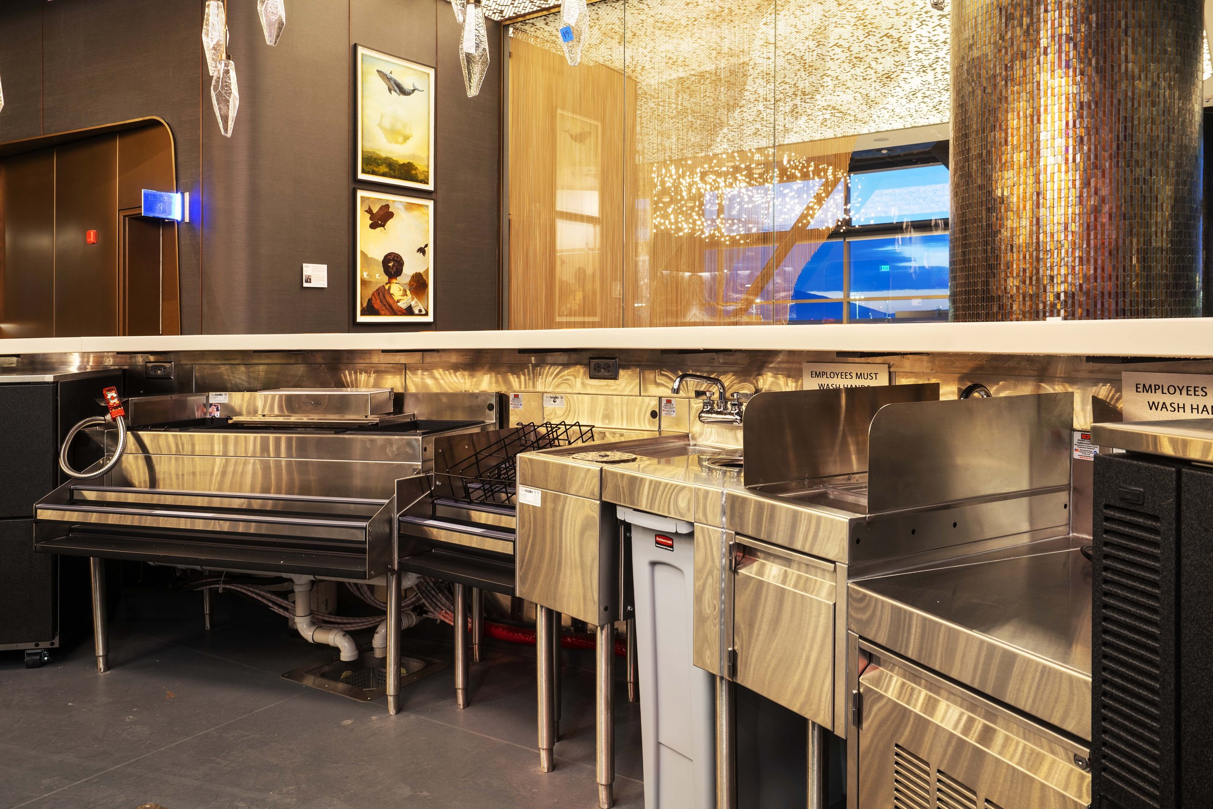 Restaurant kitchen area with stainless steel sinks and counters, signs instructing employees to wash hands, dark wall with framed artwork, reflective glass window, and decorative hanging lights.