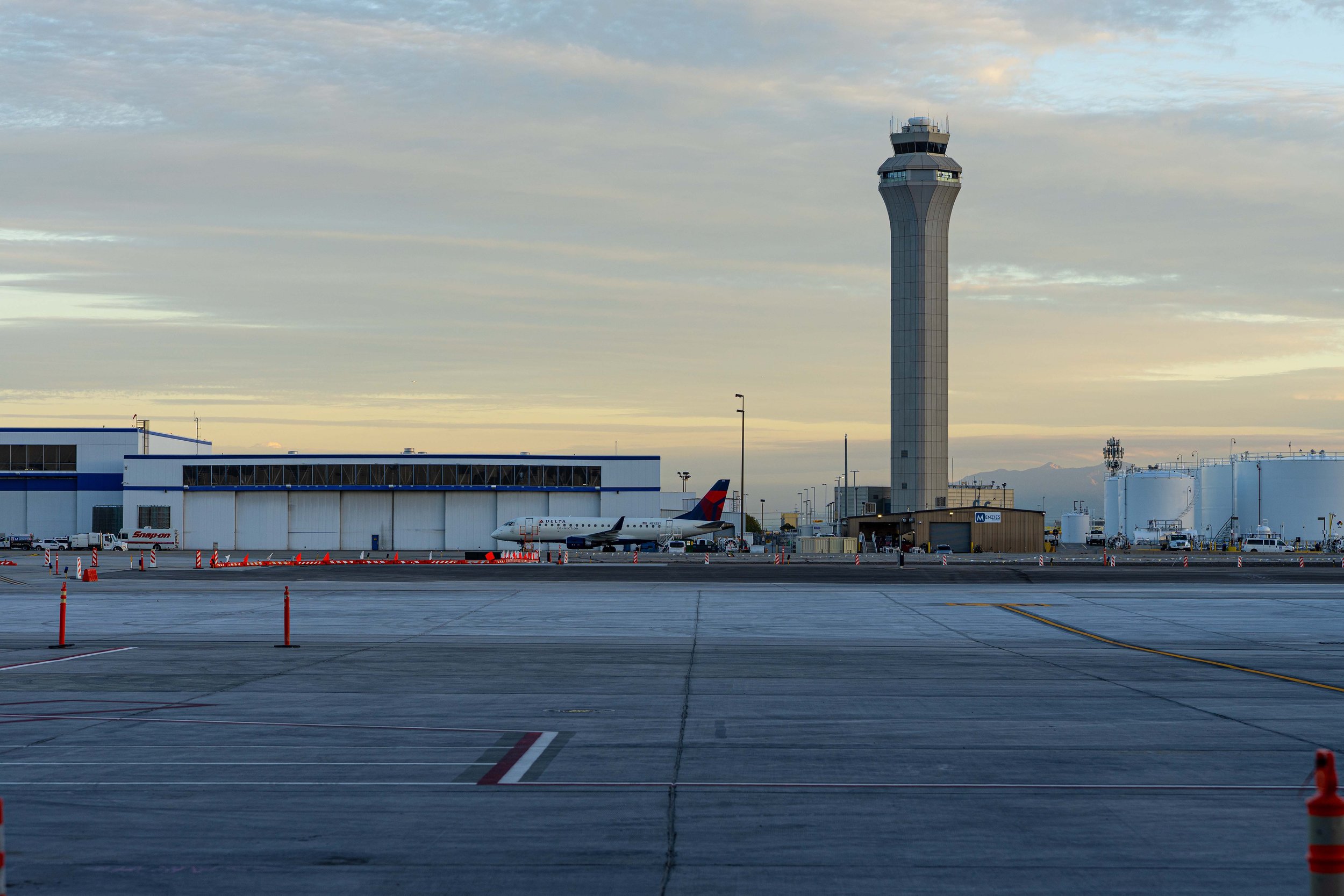 Airport tarmac with a Delta aircraft parked near a terminal, control tower in the background, and sunset sky.