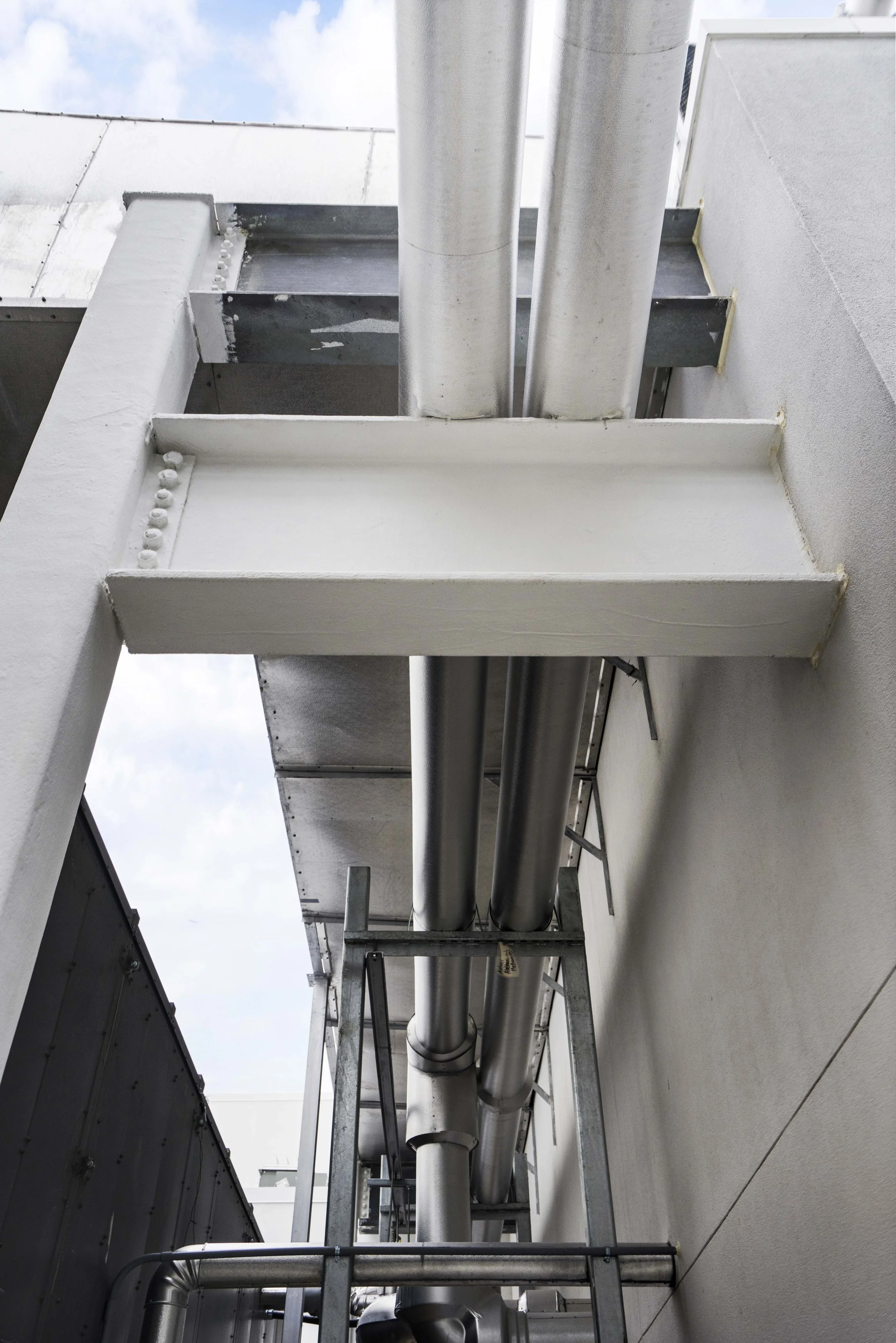 View of industrial pipes running along the side of a building, supported by metal brackets and a white painted steel beam.