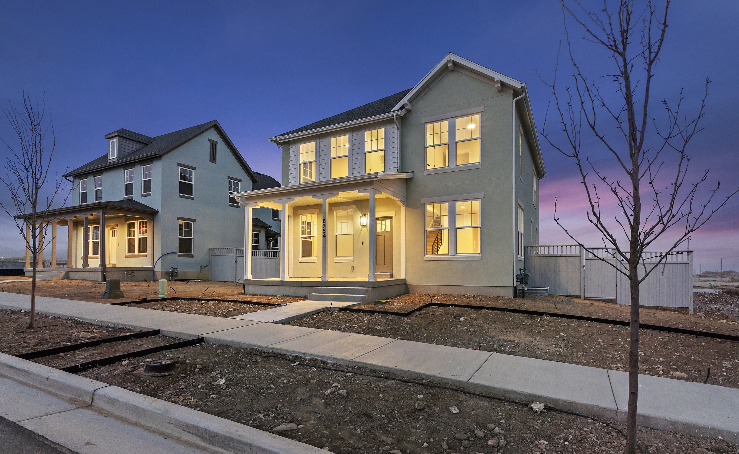Two newly constructed houses with lit interiors at dusk, featuring large windows and minimal landscaping in the foreground.