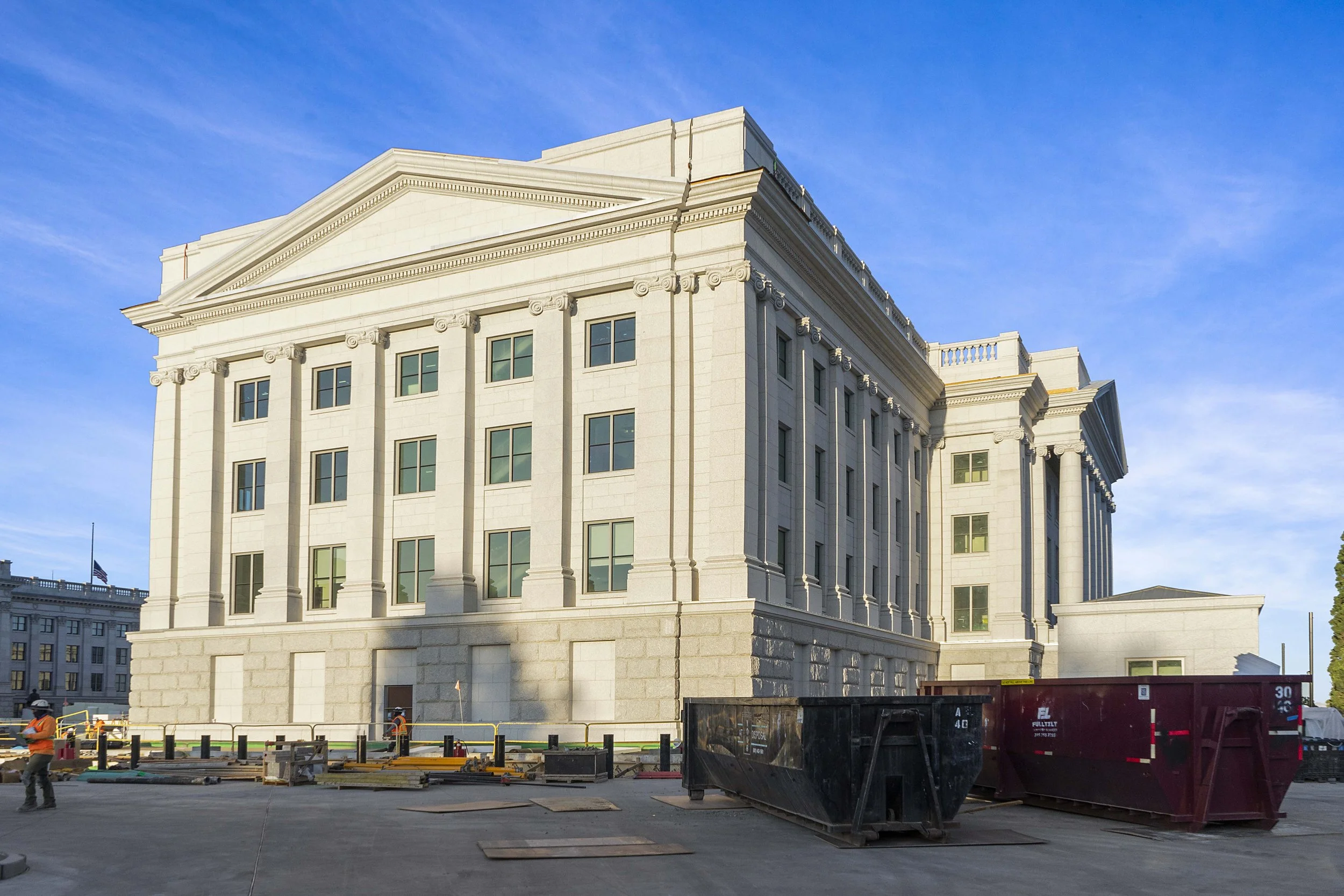 Construction site in front of a neoclassical building with columns and decorative details, under a clear blue sky.