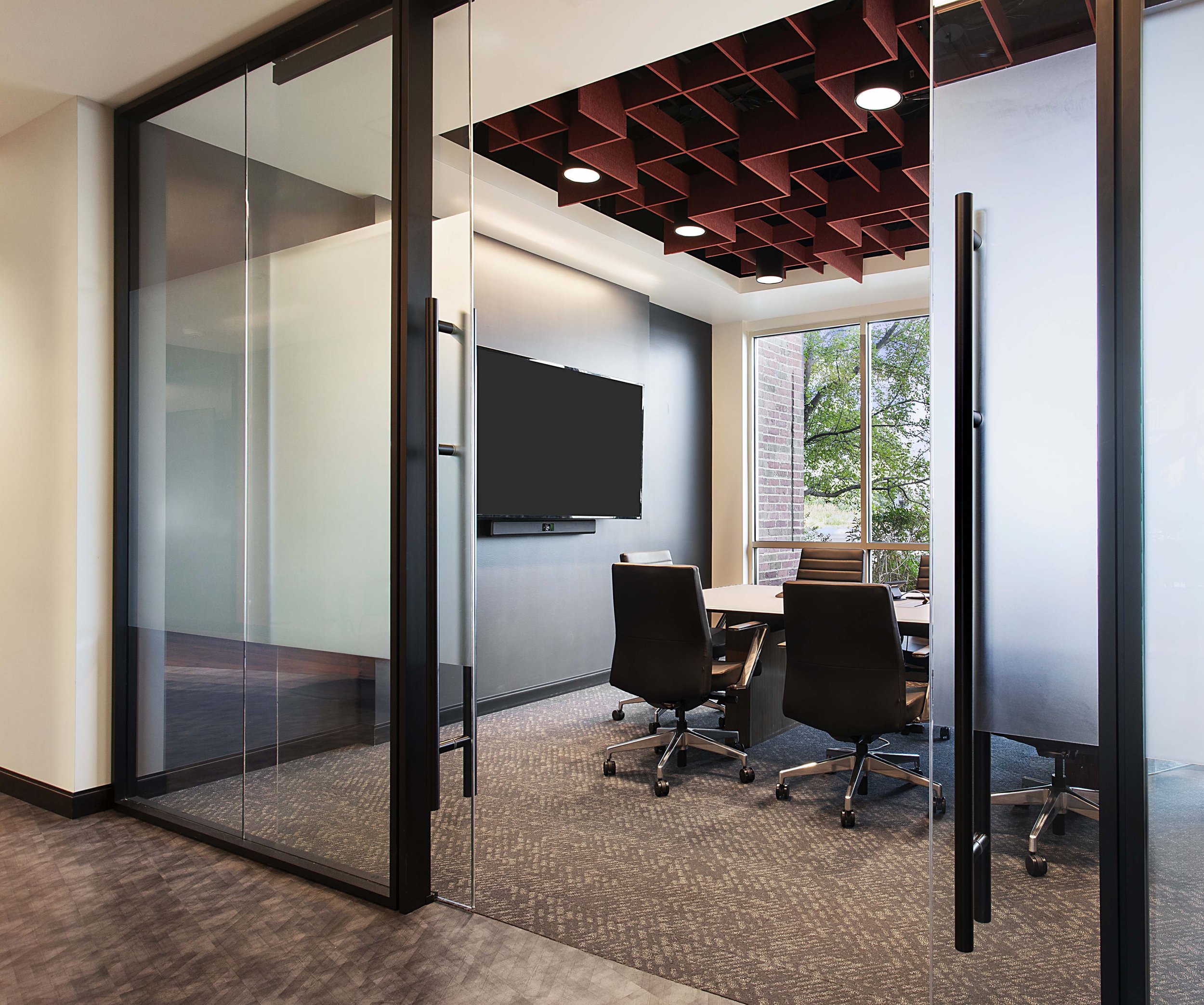 Modern conference room with glass walls, a large TV, and a table with comfortable chairs, next to a window with green trees outside.