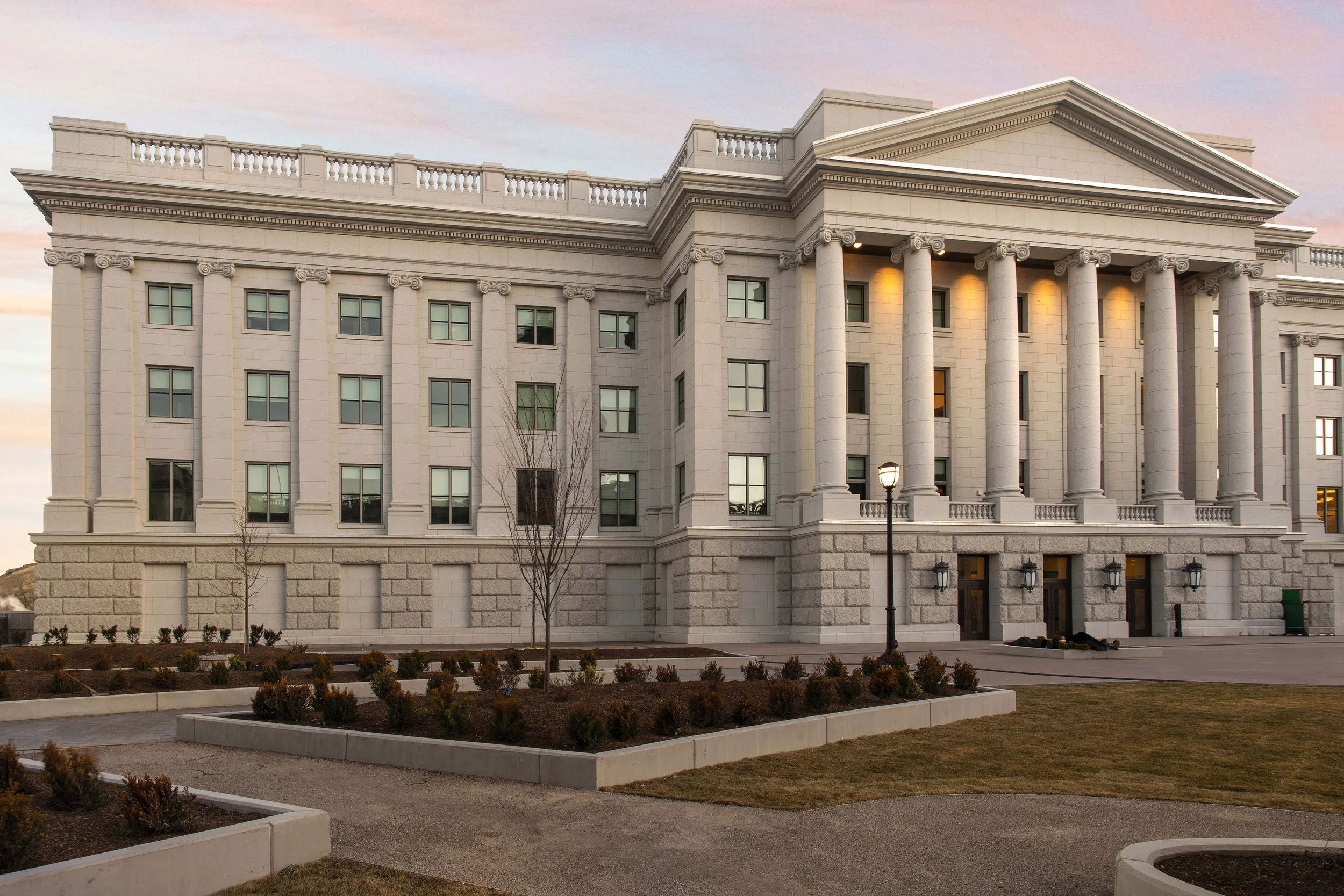 A neoclassical building with tall columns and a triangular pediment, illuminated by yellow lights, at sunset. The building has multiple windows and a landscaped area with small bushes and pathways in front.