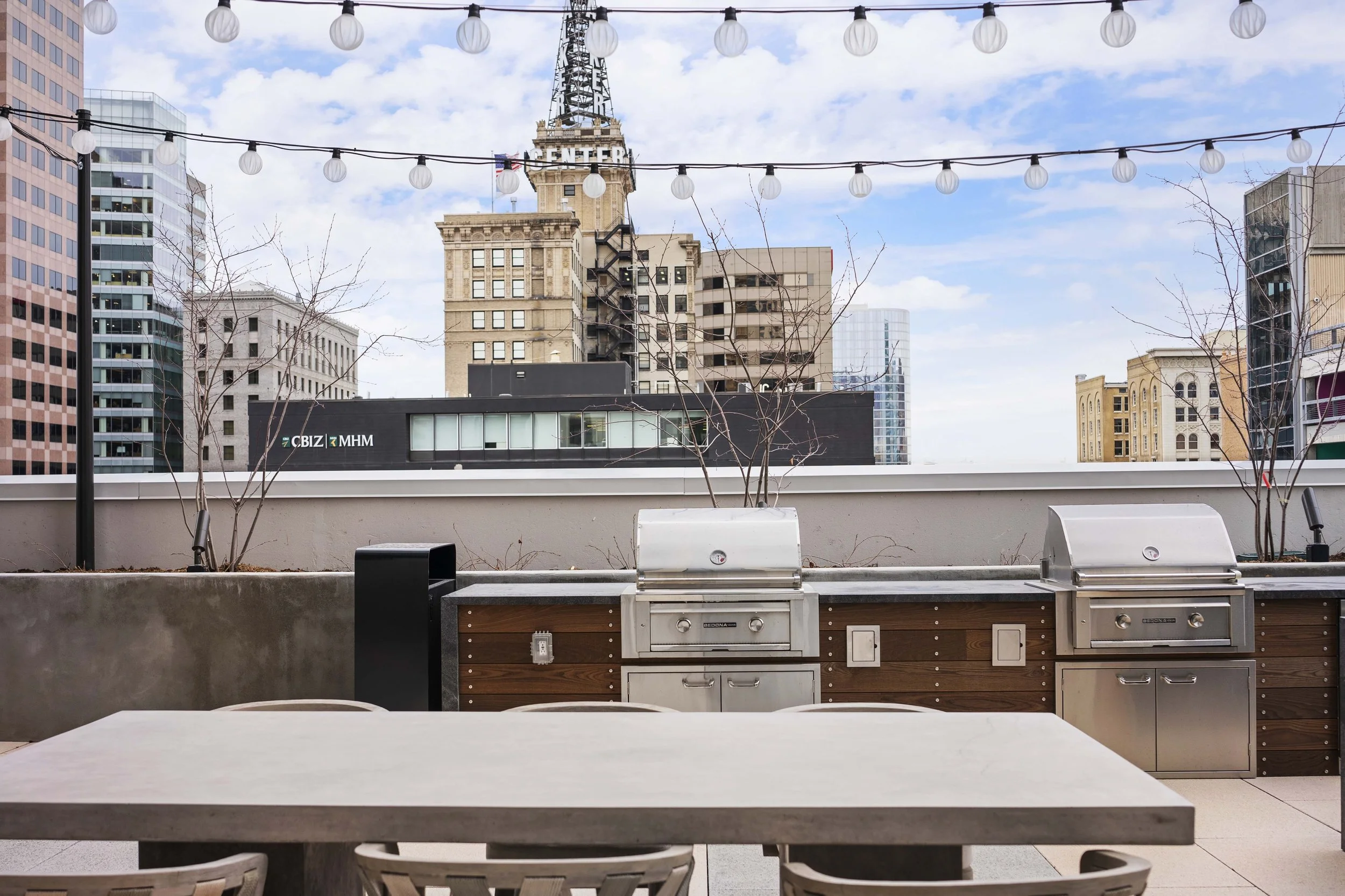 Rooftop outdoor kitchen with two grills, dining table, and city skyline with buildings under a partly cloudy sky.