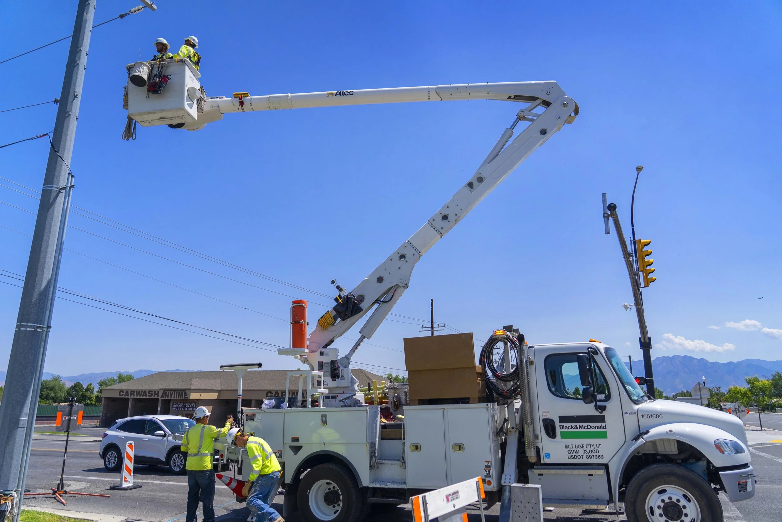 Utility workers in safety gear using a bucket lift on a truck to work on power lines at a pole, with a traffic light and a car in the background under a clear blue sky.