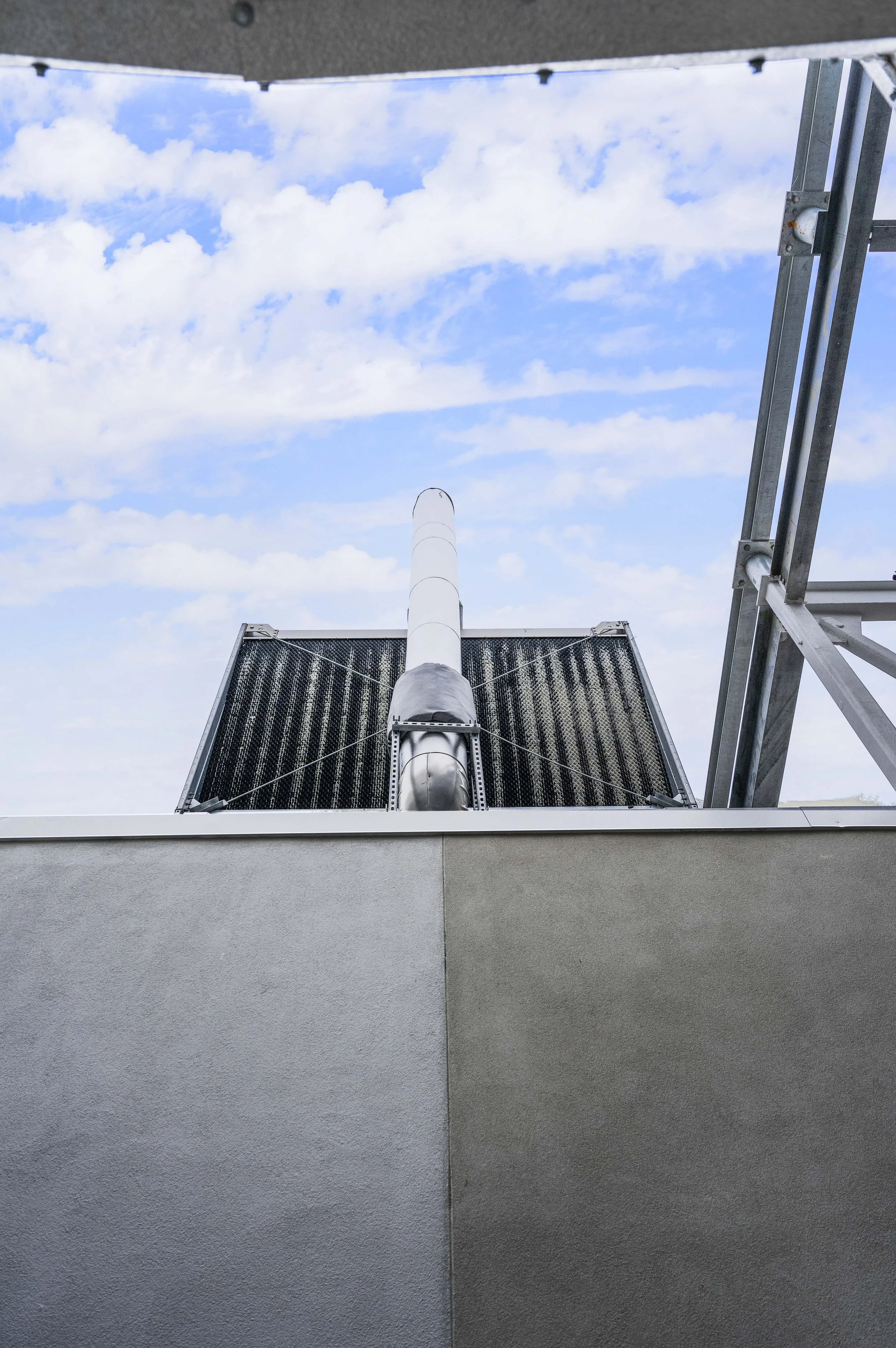 View of an industrial exhaust pipe and solar panels on a roof, with a partly cloudy sky in the background.