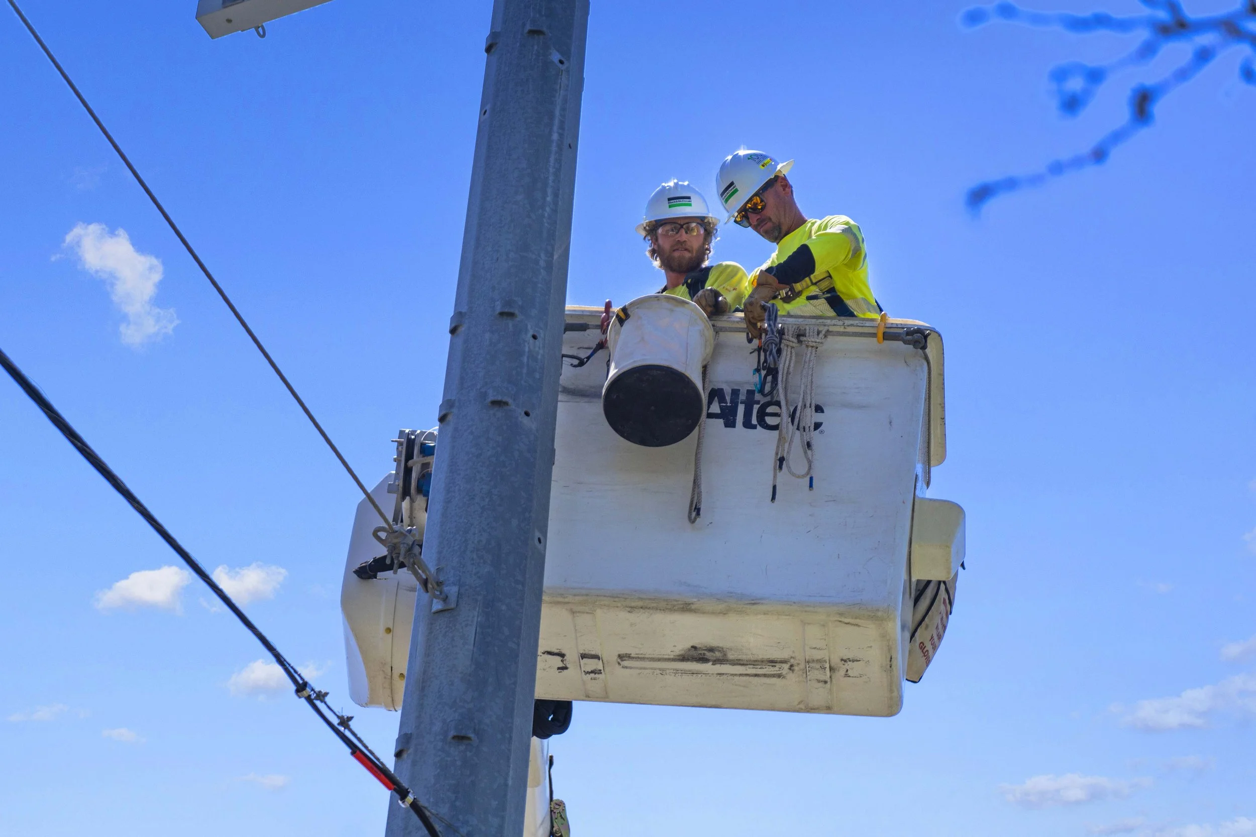 Two utility workers in safety gear and helmets working in a bucket lift on a utility pole against a blue sky with some clouds.