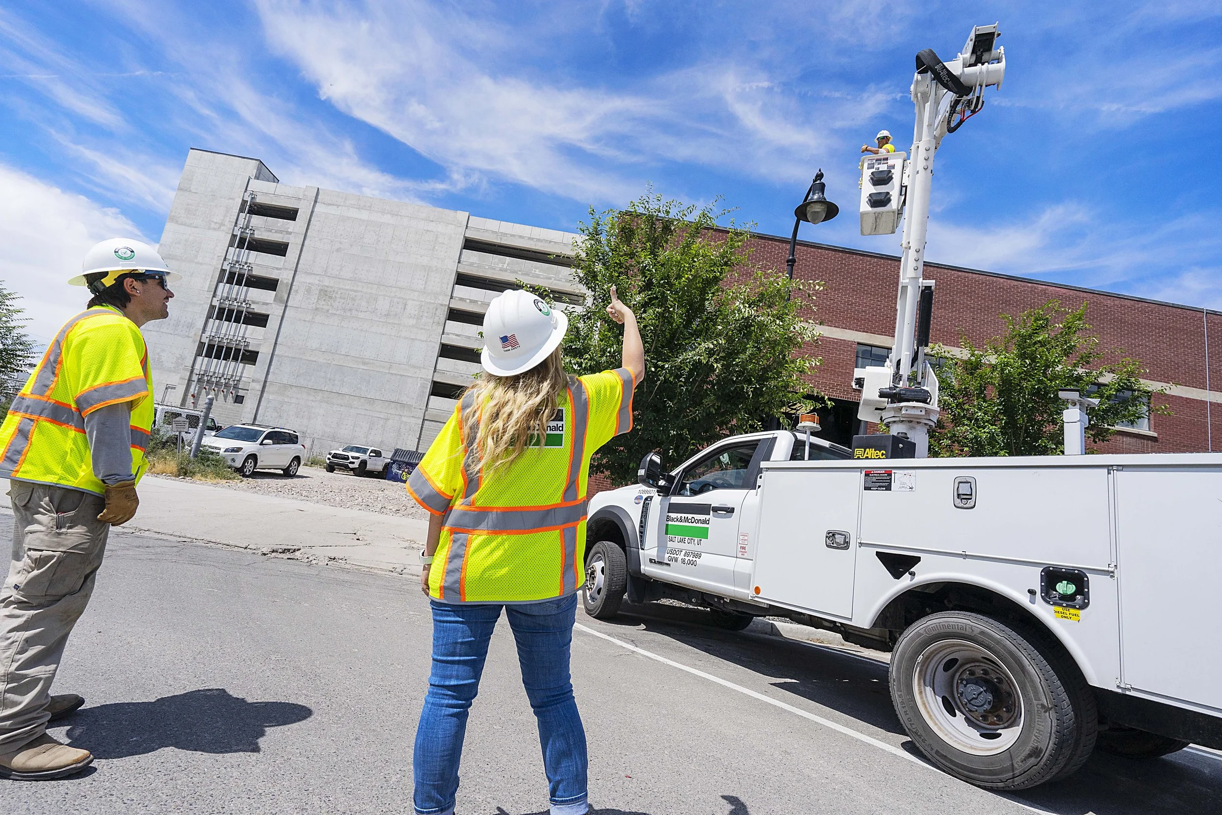 Construction workers in safety vests and helmets using a bucket truck to work on traffic signals on a city street under a blue sky.