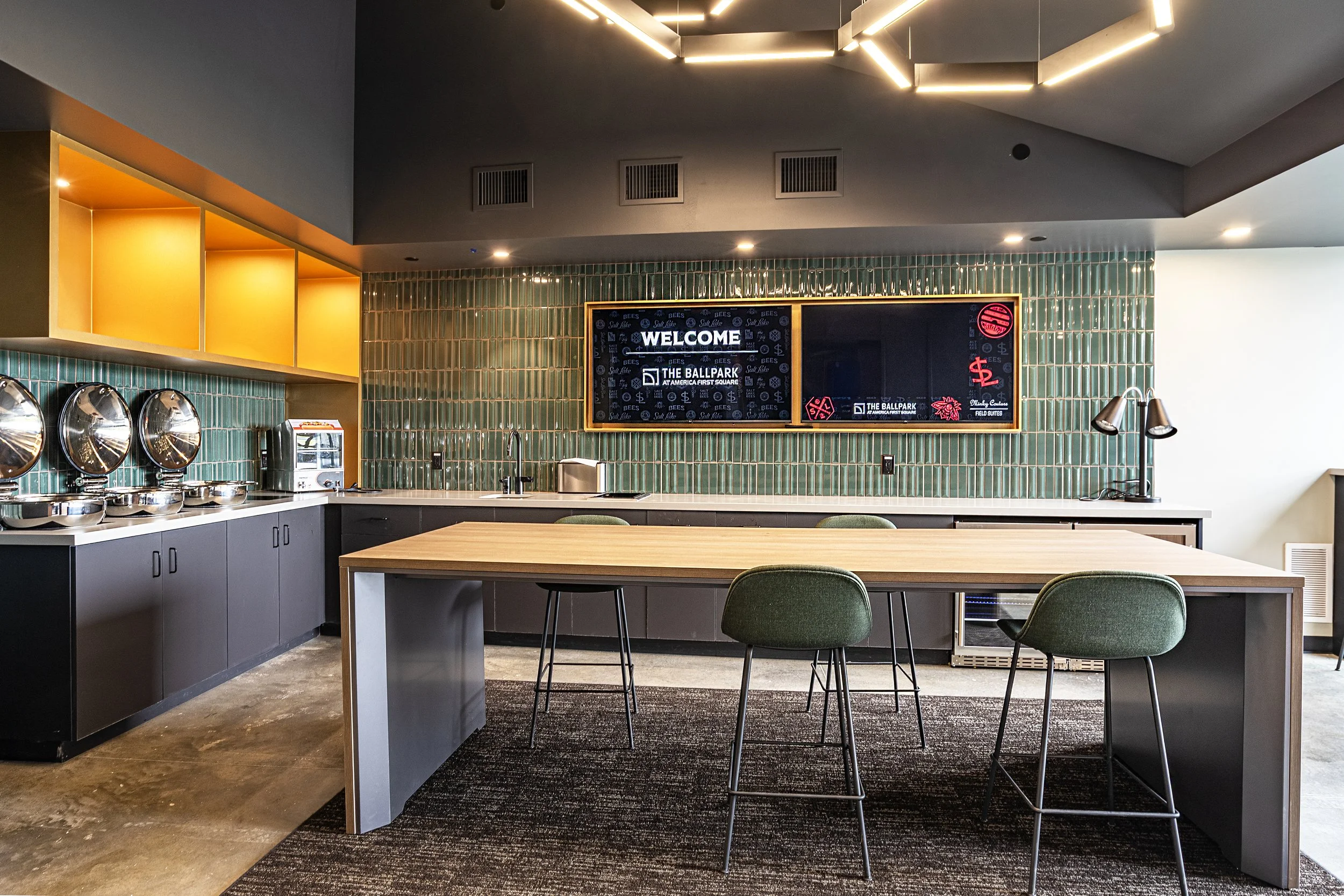 Modern conference room or kitchenette with a large wooden table surrounded by green chairs, green tiled wall with electronic displays, countertop with coffee machines and equipment, overhead lighting, and yellow wall shelves.