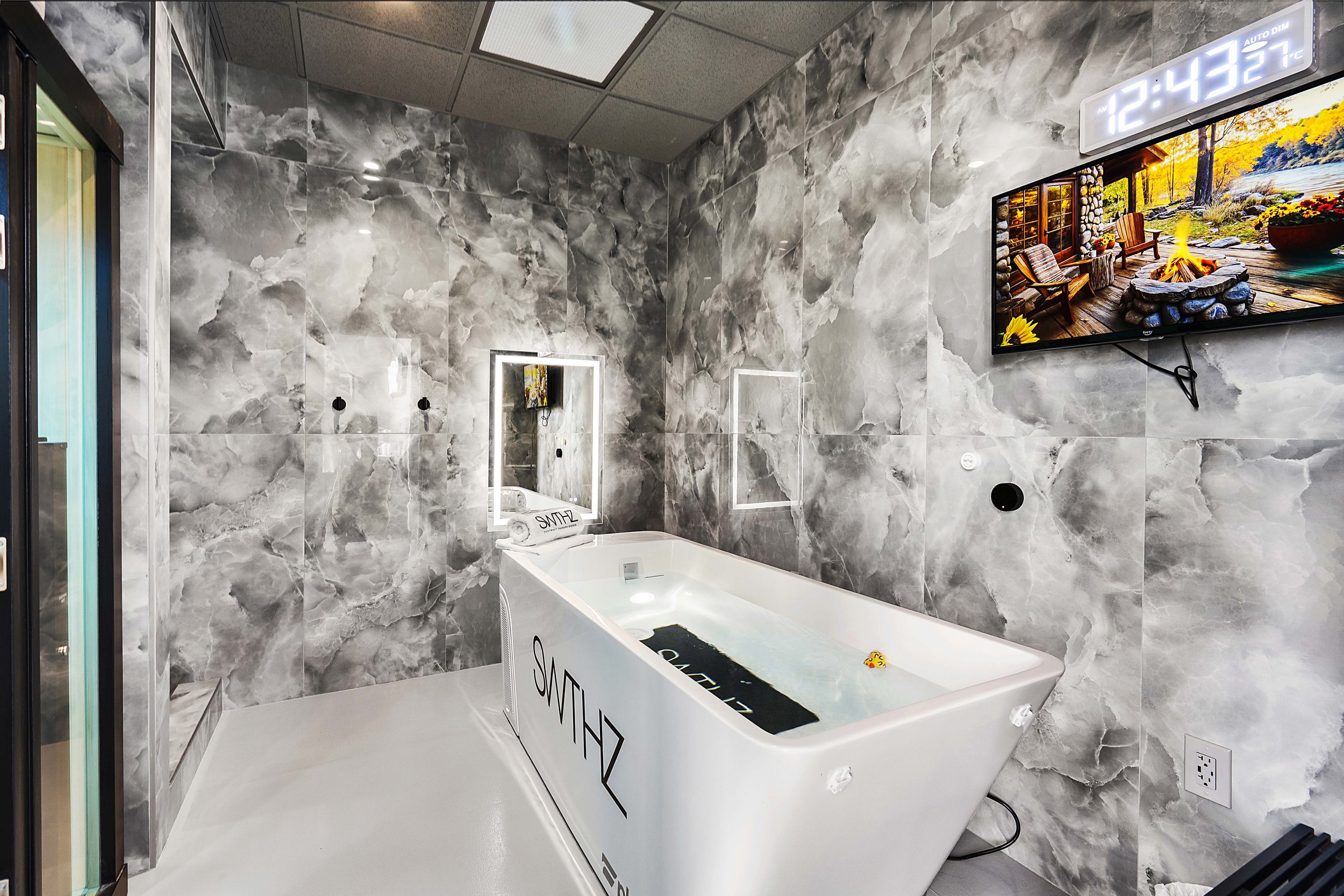 Modern indoor spa room with gray marble walls and white floor, featuring a white rectangular hot tub and a wall-mounted TV showing an outdoor scene with a fire pit and chairs.