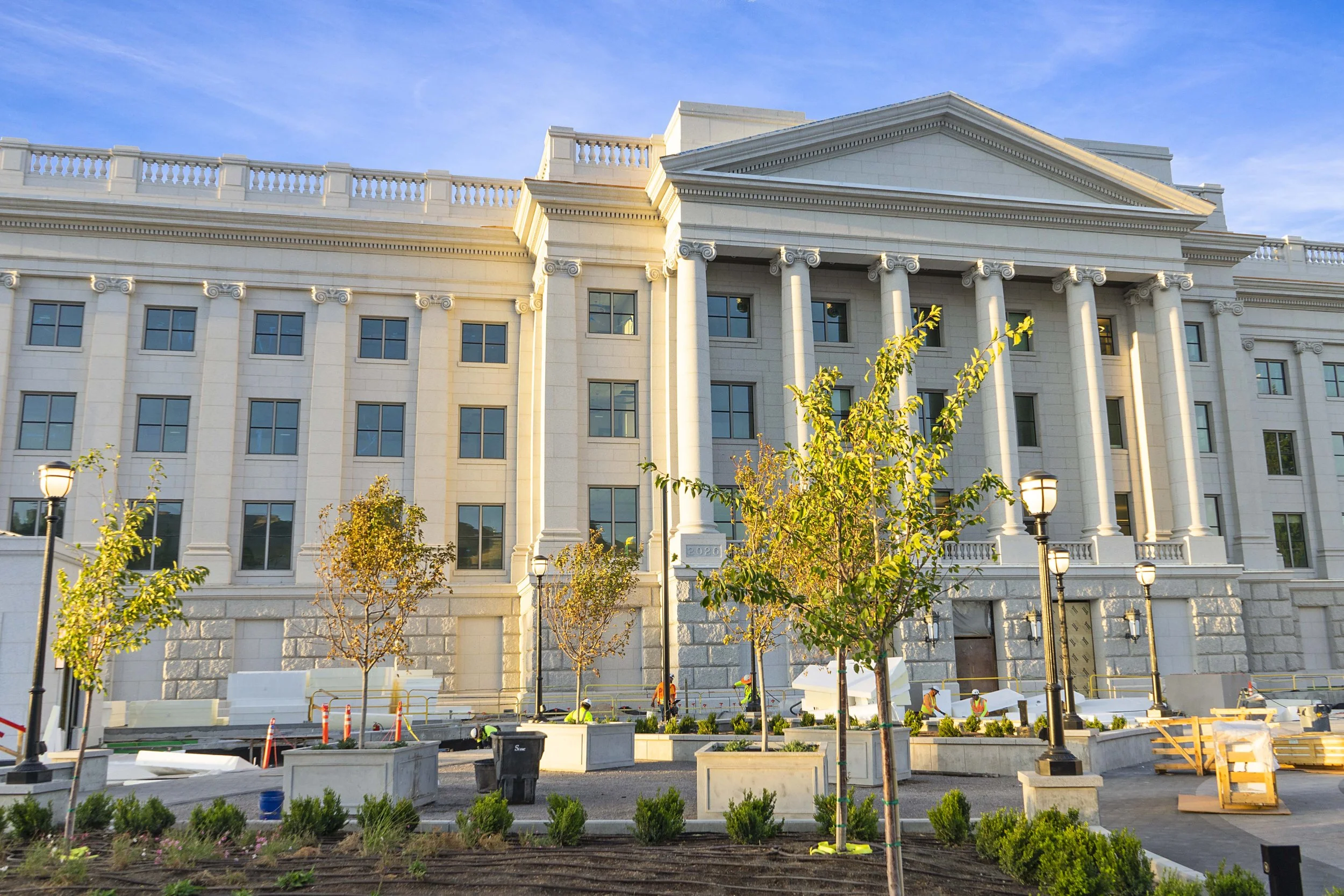 Construction workers working on a building with classical architectural features, including columns and a pediment, during daytime, with trees and street lamps in the foreground.