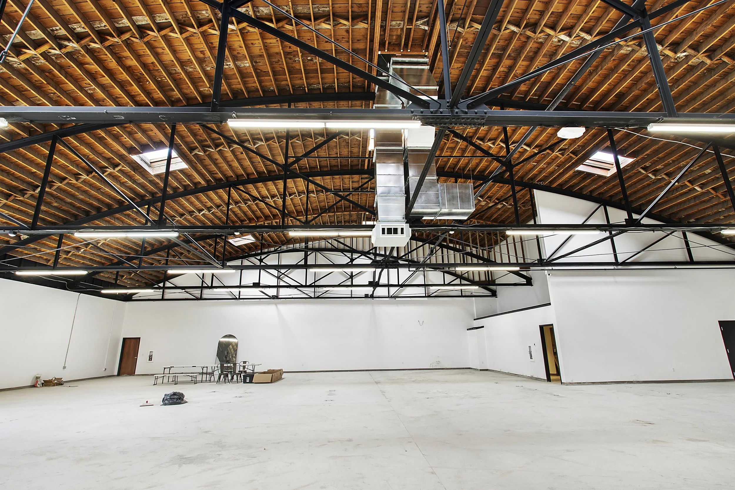 Empty warehouse or industrial space with white walls, a high wooden ceiling with exposed beams, and metal trusses. Some scattered furniture and items are present.
