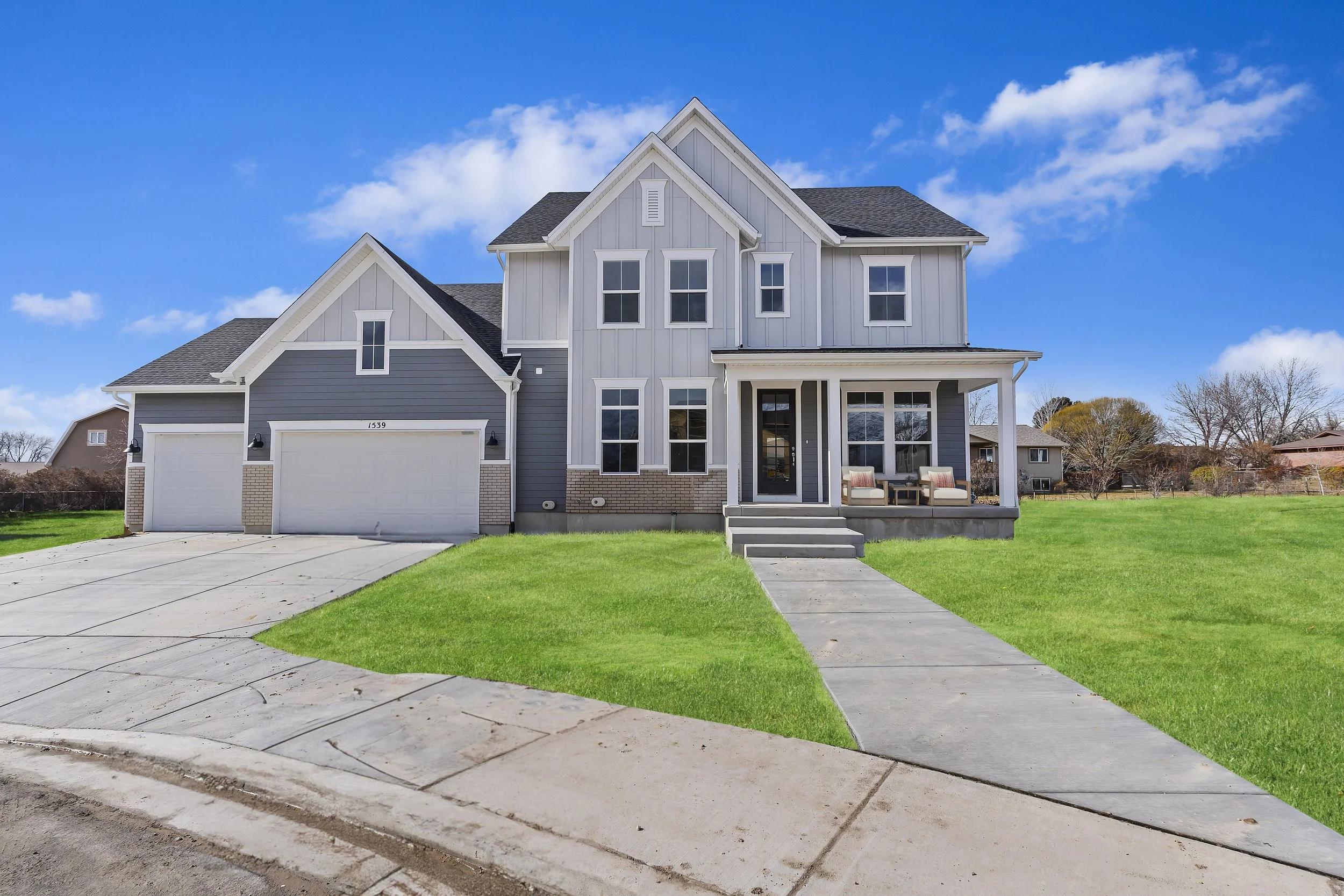 Modern gray two-story house with a front porch, double garage, and green lawn, set against a blue sky.
