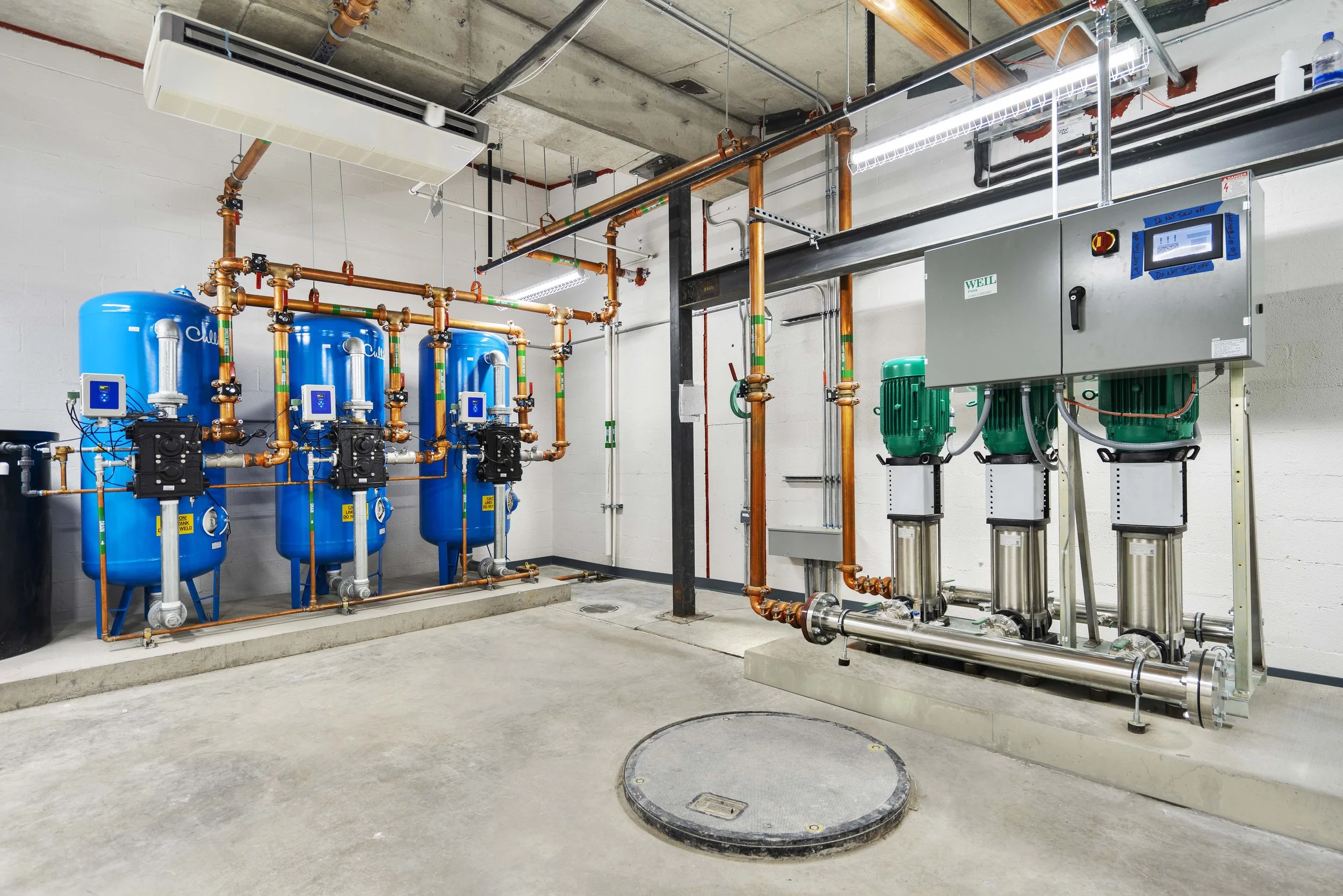 Industrial mechanical room with blue water pressure tanks, green variable frequency drives, copper pipes, wall-mounted control panels, and a concrete floor.