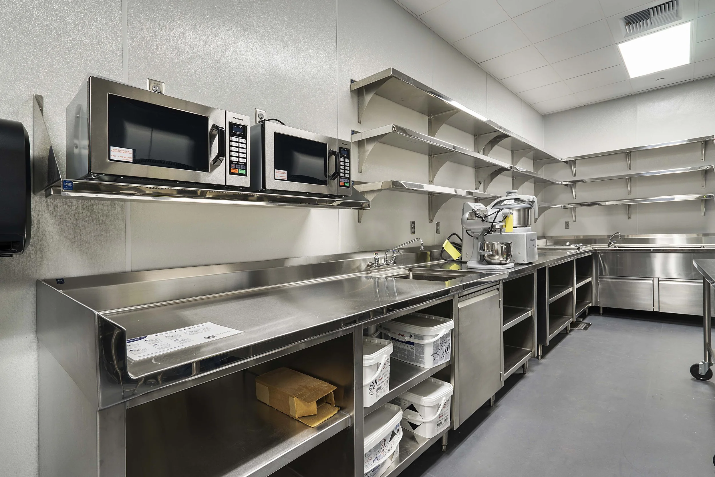 Clean commercial kitchen with stainless steel countertops, microwave ovens, industrial mixer, open shelving, and storage containers.