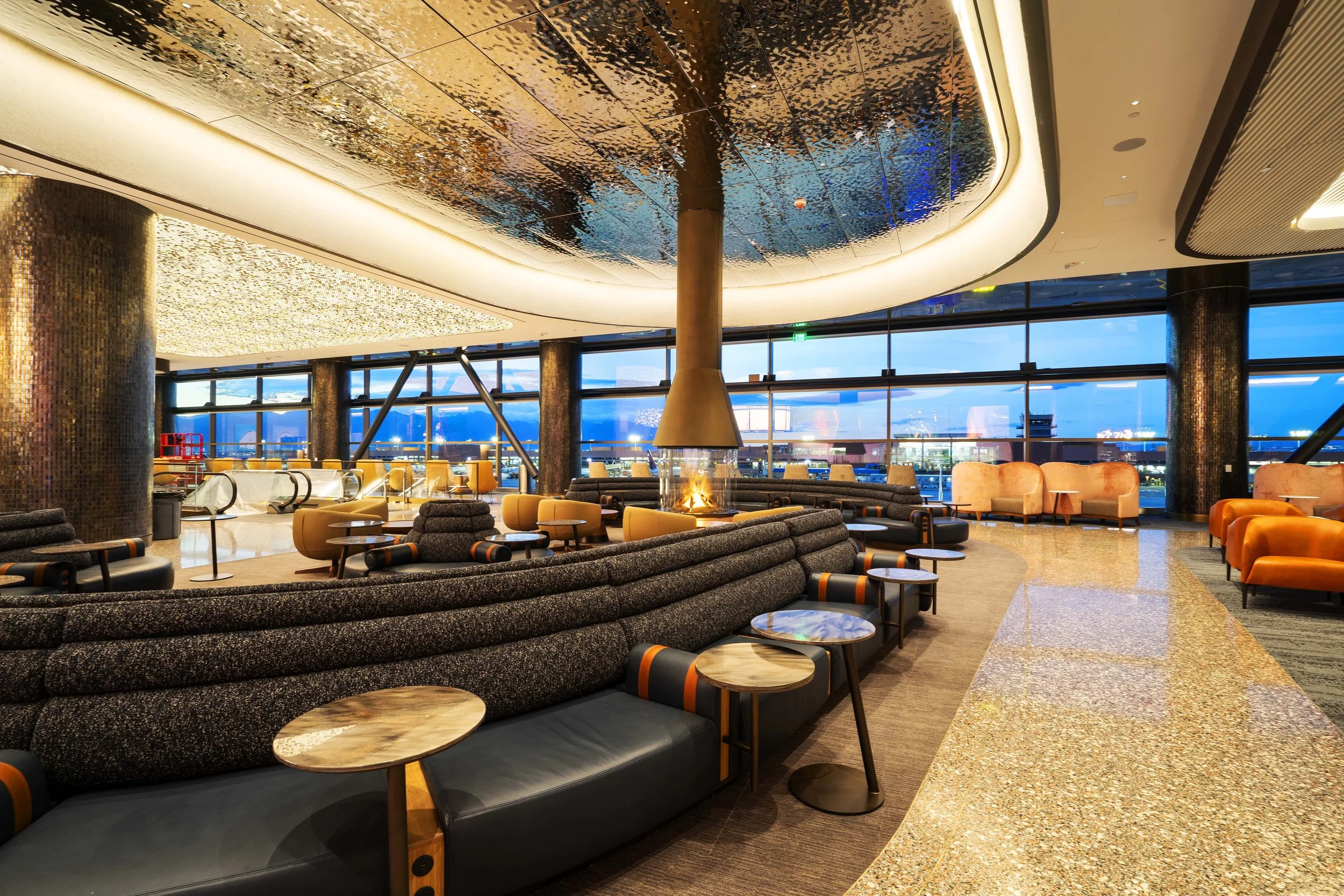 Luxury airport lounge with modern furniture, large windows overlooking the tarmac, and a fireplace in the center. The space features colorful seating and artistic ceiling decorations.