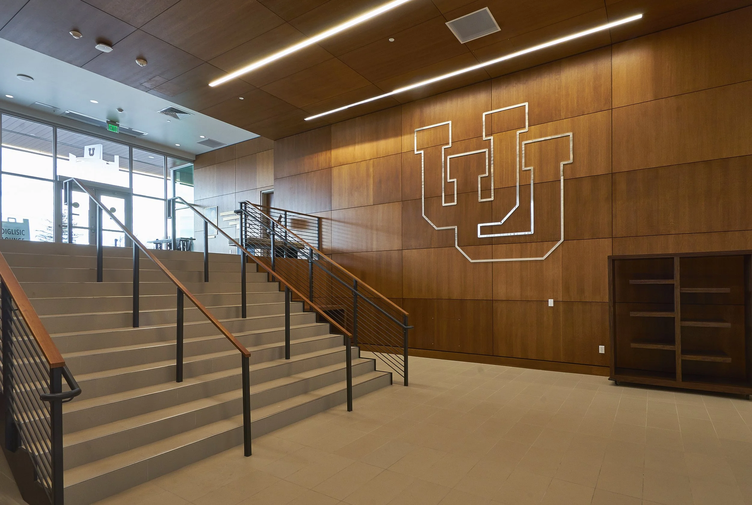Interior of a building with wooden walls, a staircase with black railing, a large illuminated University of Utah logo on the wall, and glass doors at the entrance.