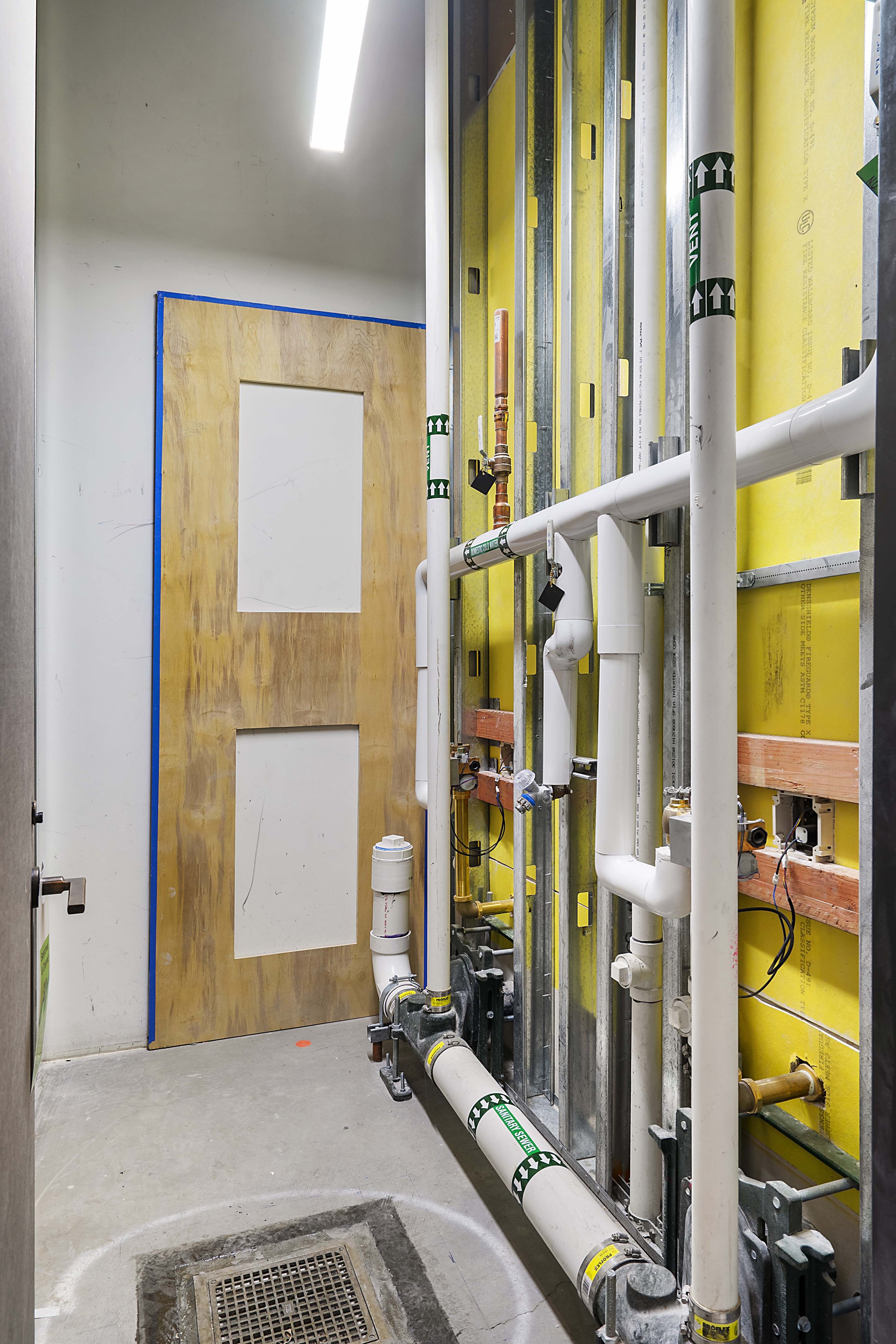 Indoor construction site with exposed plumbing, yellow insulation, a plywood door, and a floor drain.