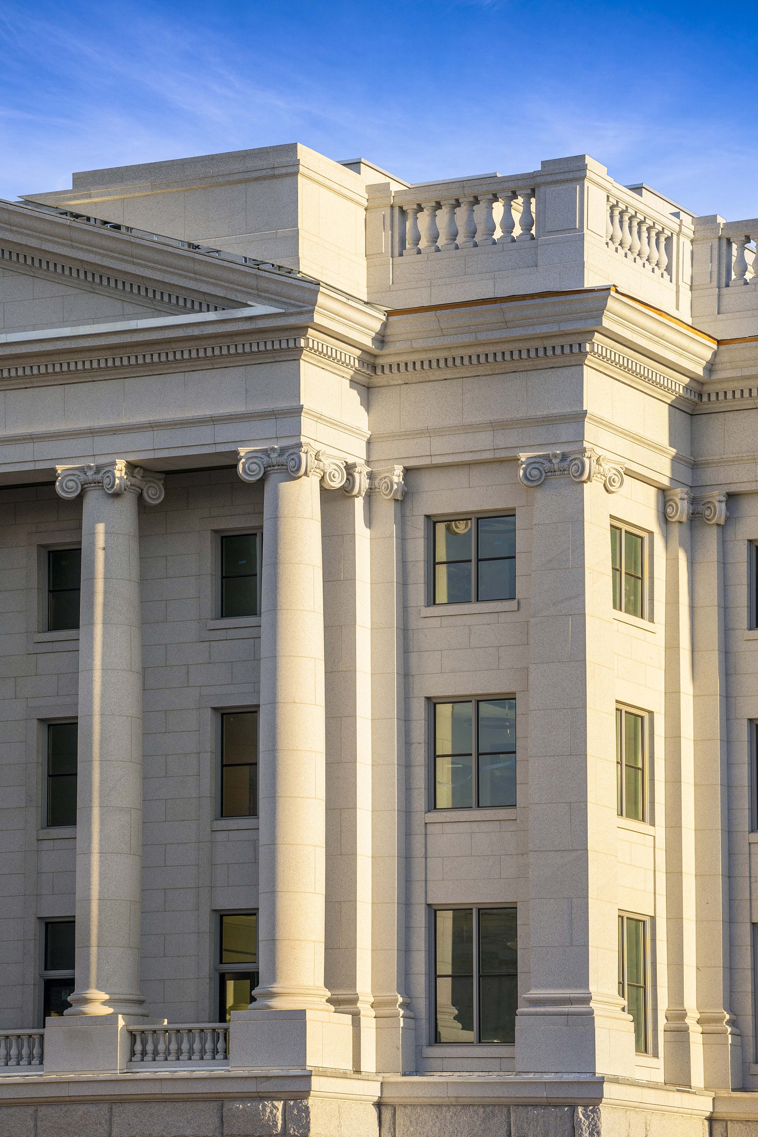 Close-up of a classical style building with large columns, ornate moldings, and a decorative balustrade, bathed in sunlight against a blue sky.