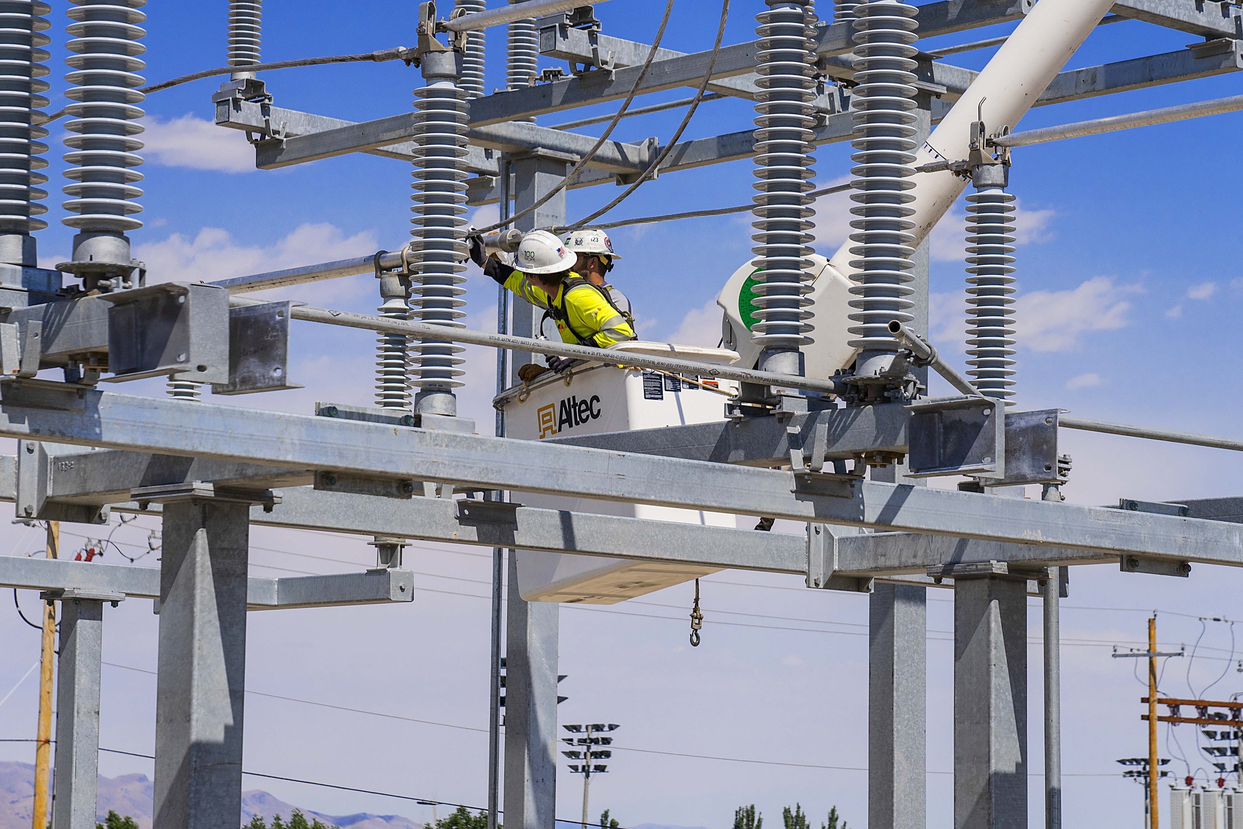 Two workers in safety gear repairing electrical power lines on a high-voltage transmission tower against a blue sky.