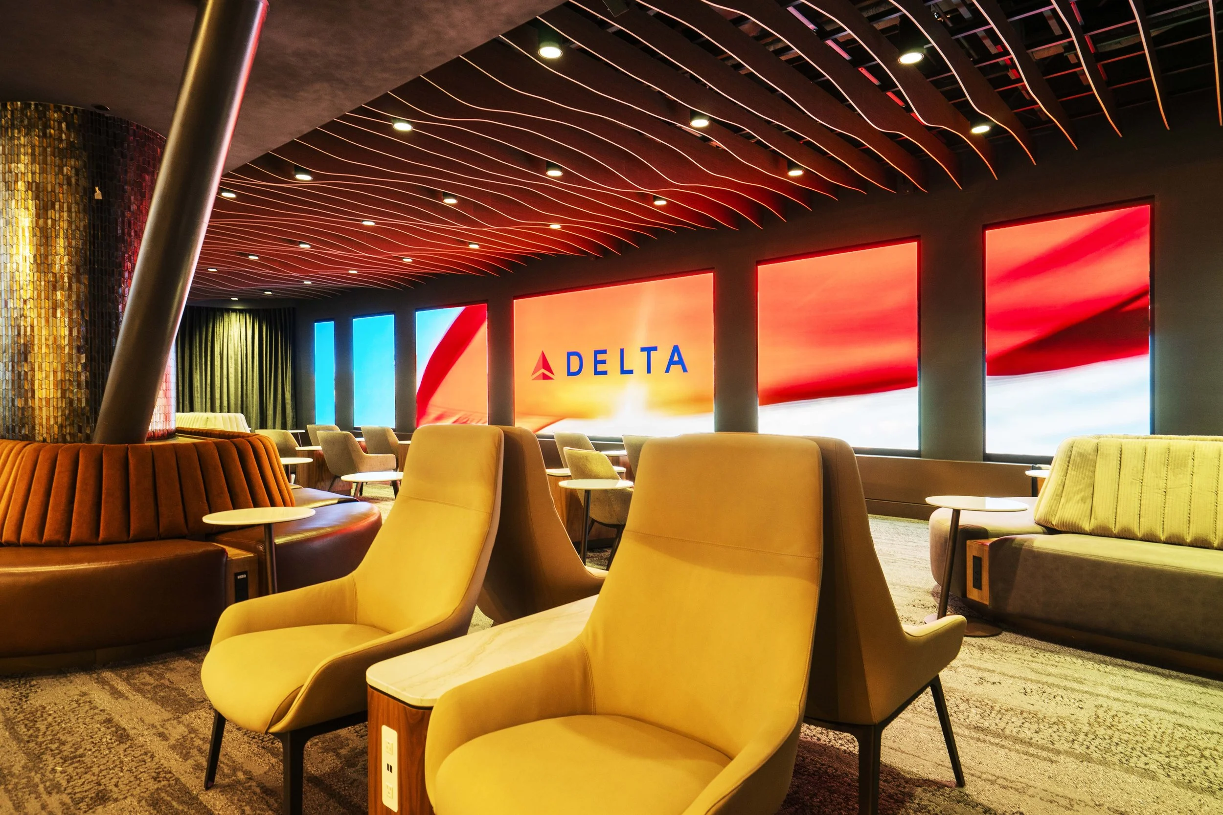 Modern airport lounge with yellow and beige chairs, illuminated digital displays on the wall, including a Delta Airlines advertisement, and colorful LED lighting on the ceiling.