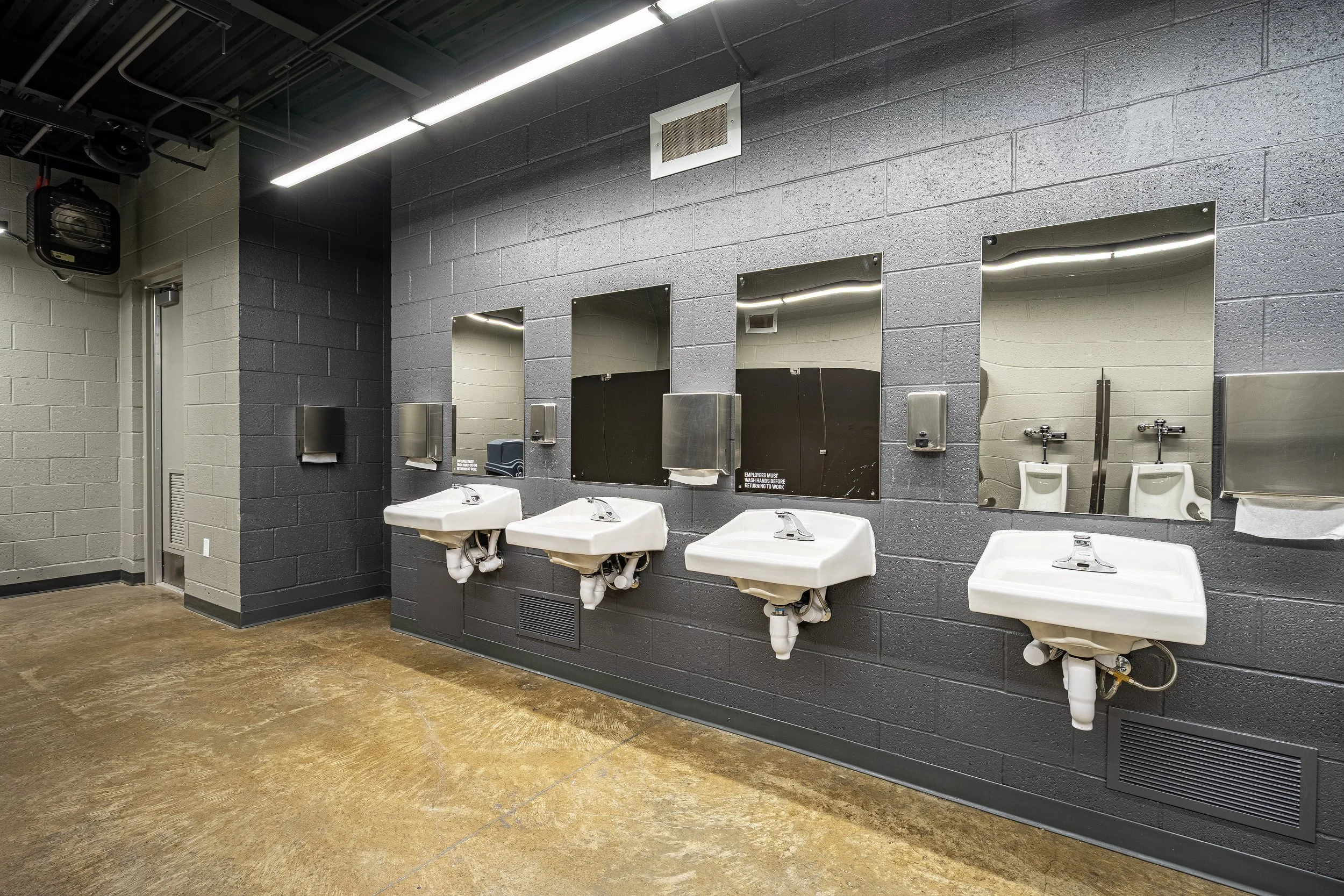 Public restroom with four sinks, four mirrors, and three soap dispensers mounted on a dark gray brick wall.