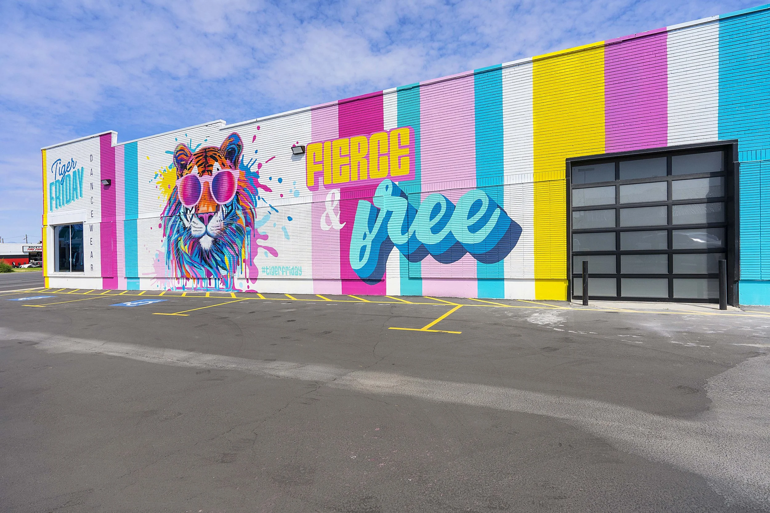 Colorful mural on building wall advertising Fierce & Free, featuring a tiger with pink sunglasses and vibrant paint splashes, with blue, pink, yellow, and white vertical stripes in the background.