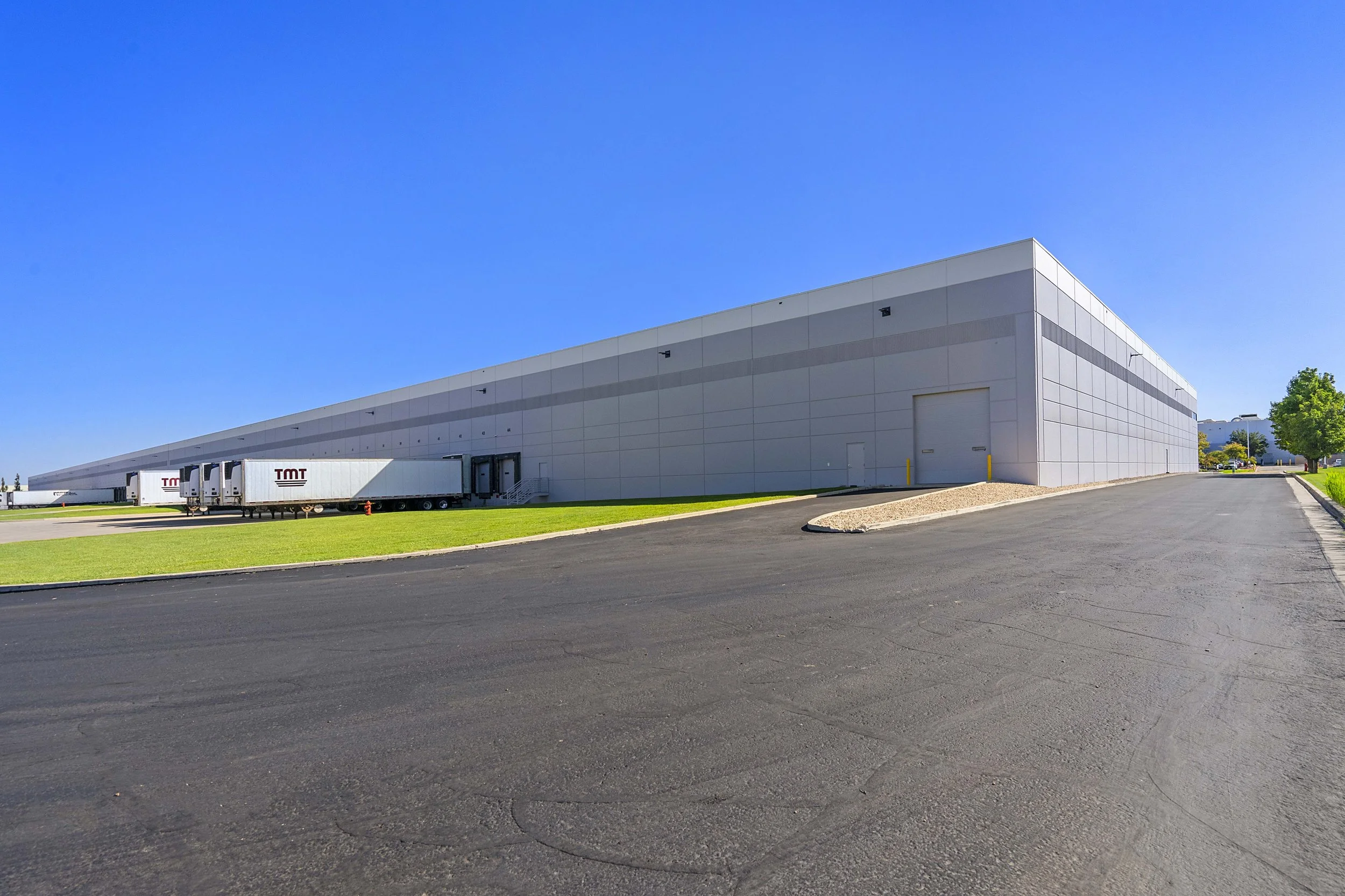 Large modern warehouse with loading docks, trucks, and trucks with TMT branding parked outside, clear blue sky, and a paved road in the foreground.