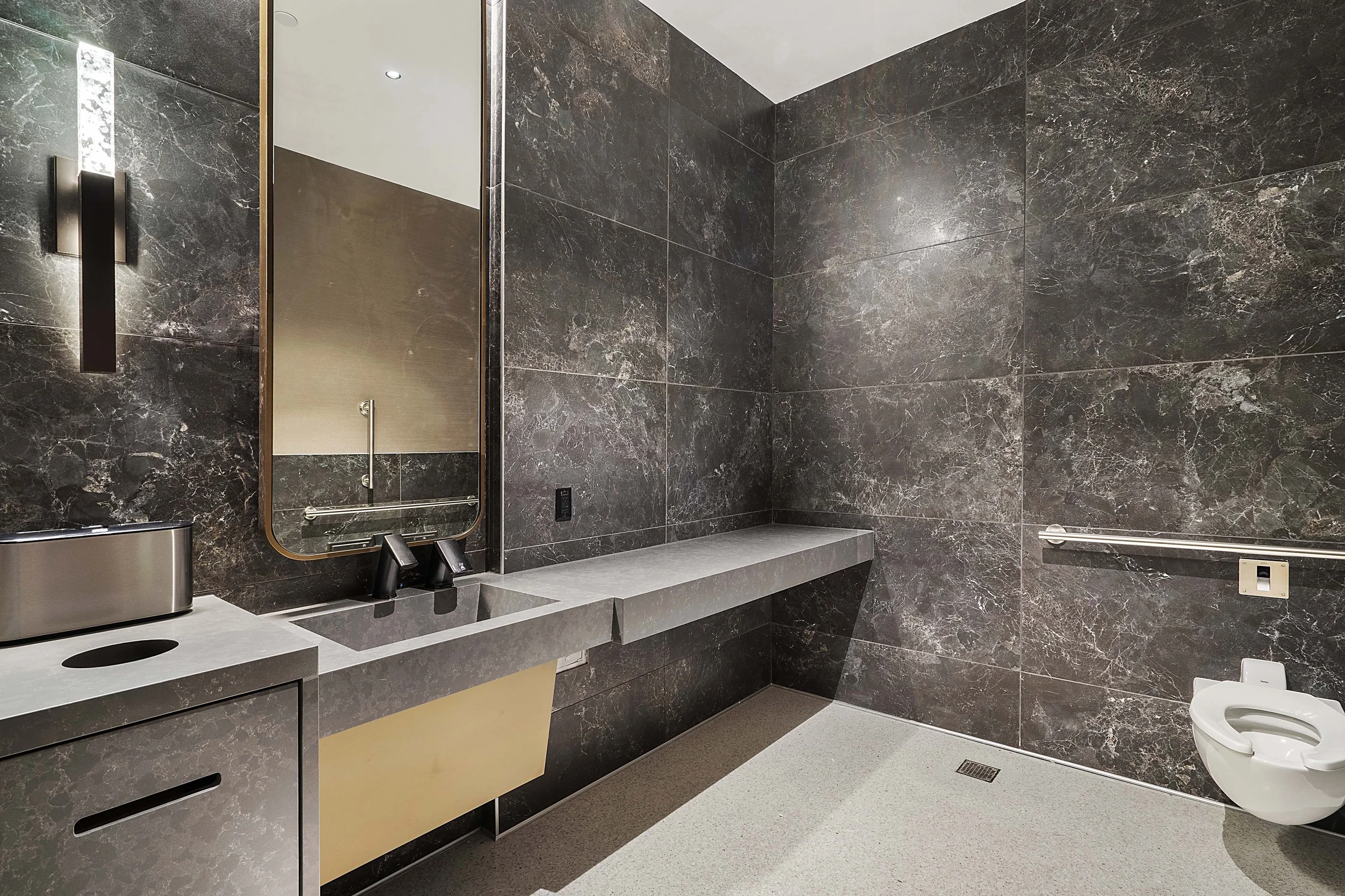 Modern bathroom with dark marble walls, a large mirror, a countertop with a sink, a tissue box, and a wall-mounted toilet.