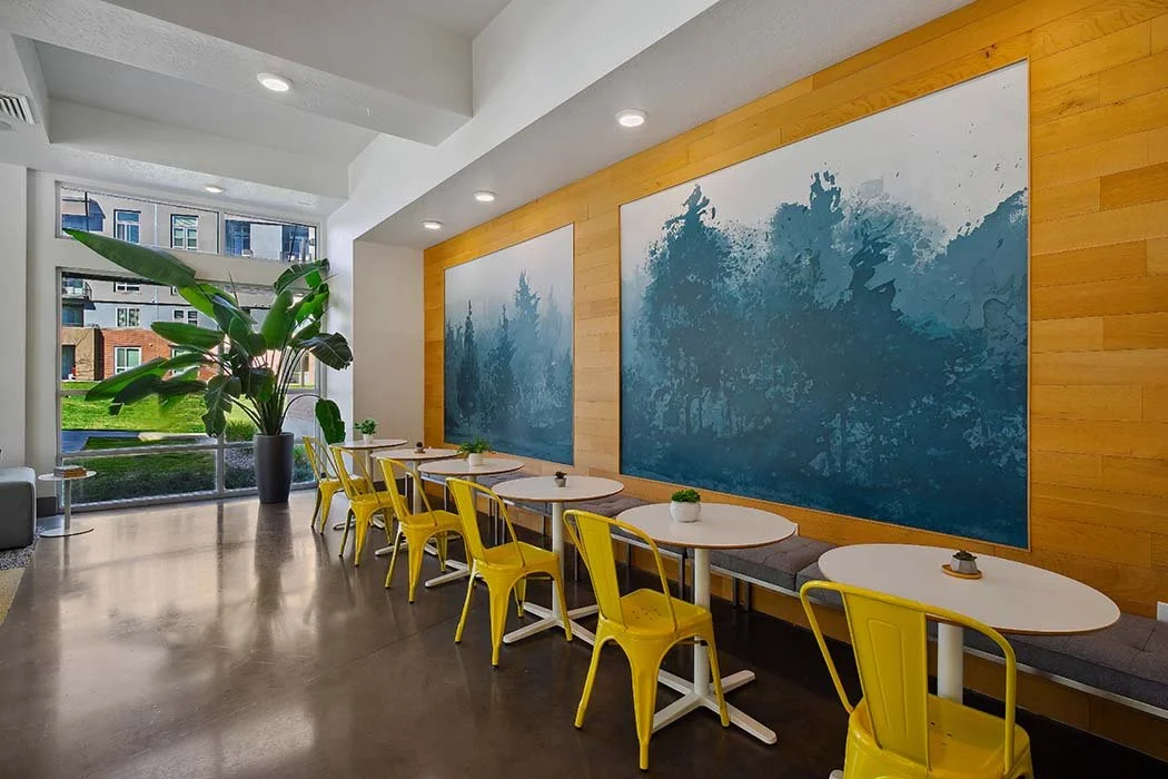 Modern cafe interior with yellow chairs, white round tables, large window with outdoor view, wooden wall with two large blue artworks, and a tall green plant in a gray pot.
