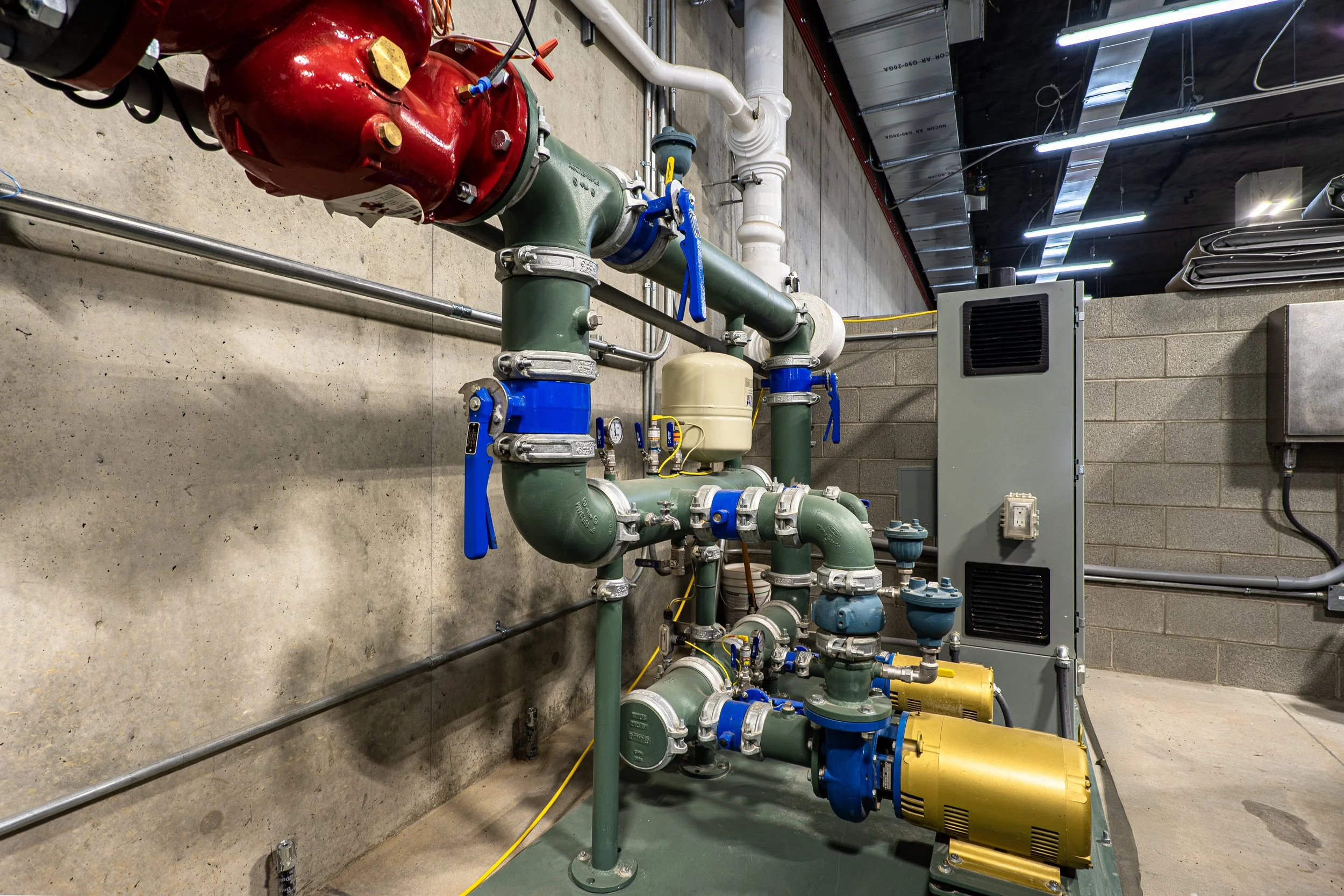 Industrial piping system with green pipes, blue valves, yellow pumps, and other equipment installed on a concrete floor near a brick wall in a utility room.