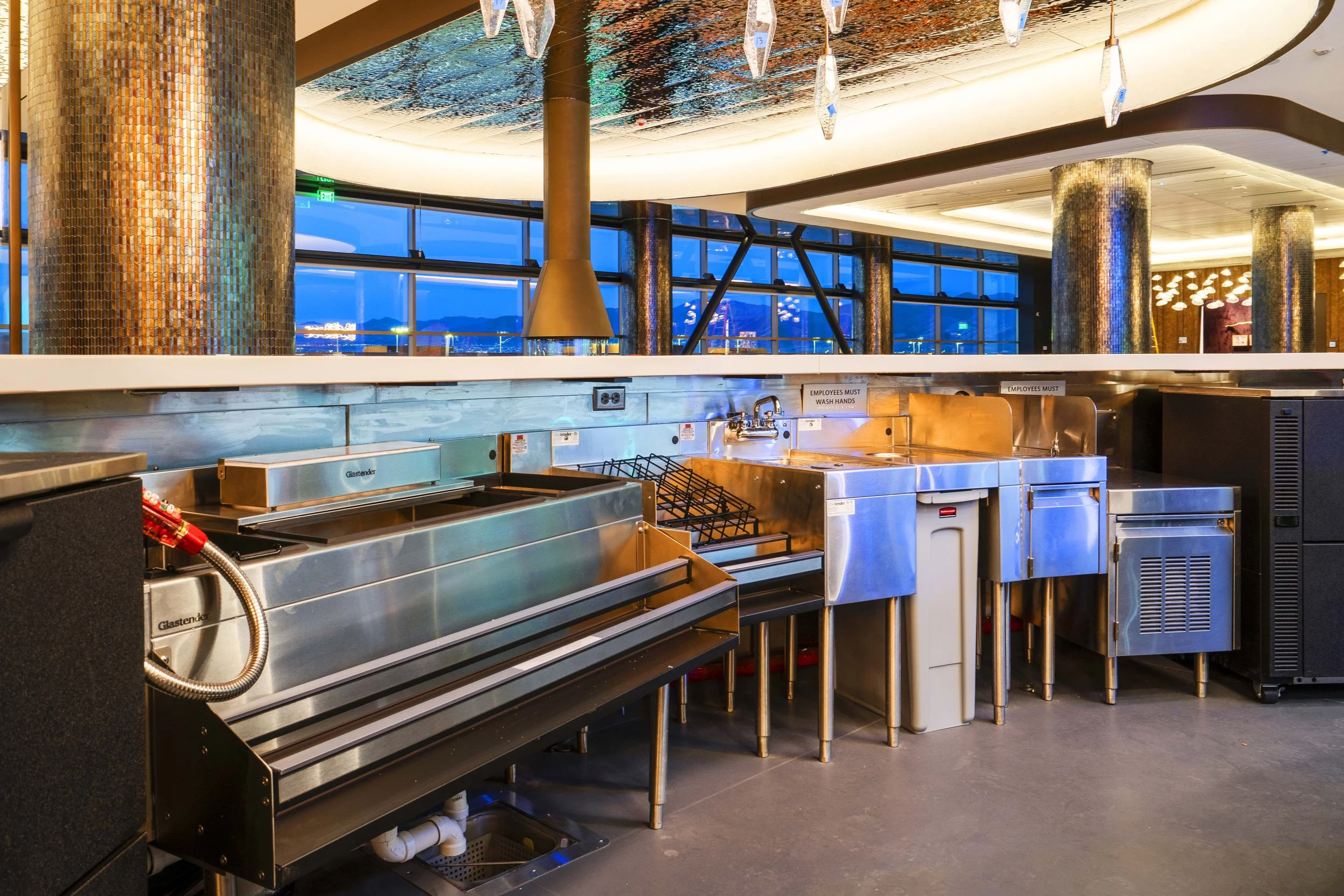 Commercial kitchen area with stainless steel appliances, large windows with city view, modern decor, and ambient lighting.