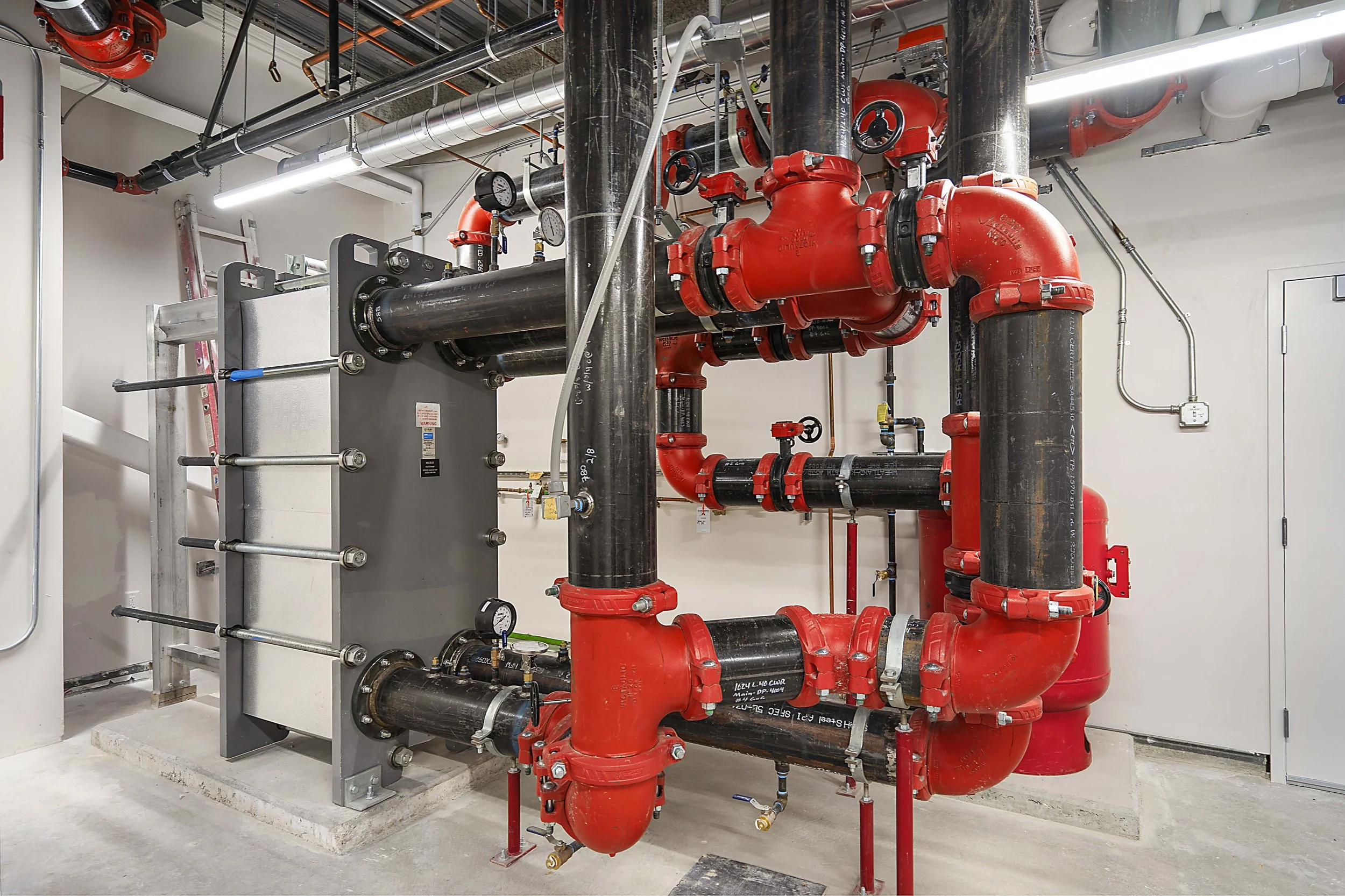 Industrial boiler room with black and red pipes, gauges, and a gray heat exchanger.